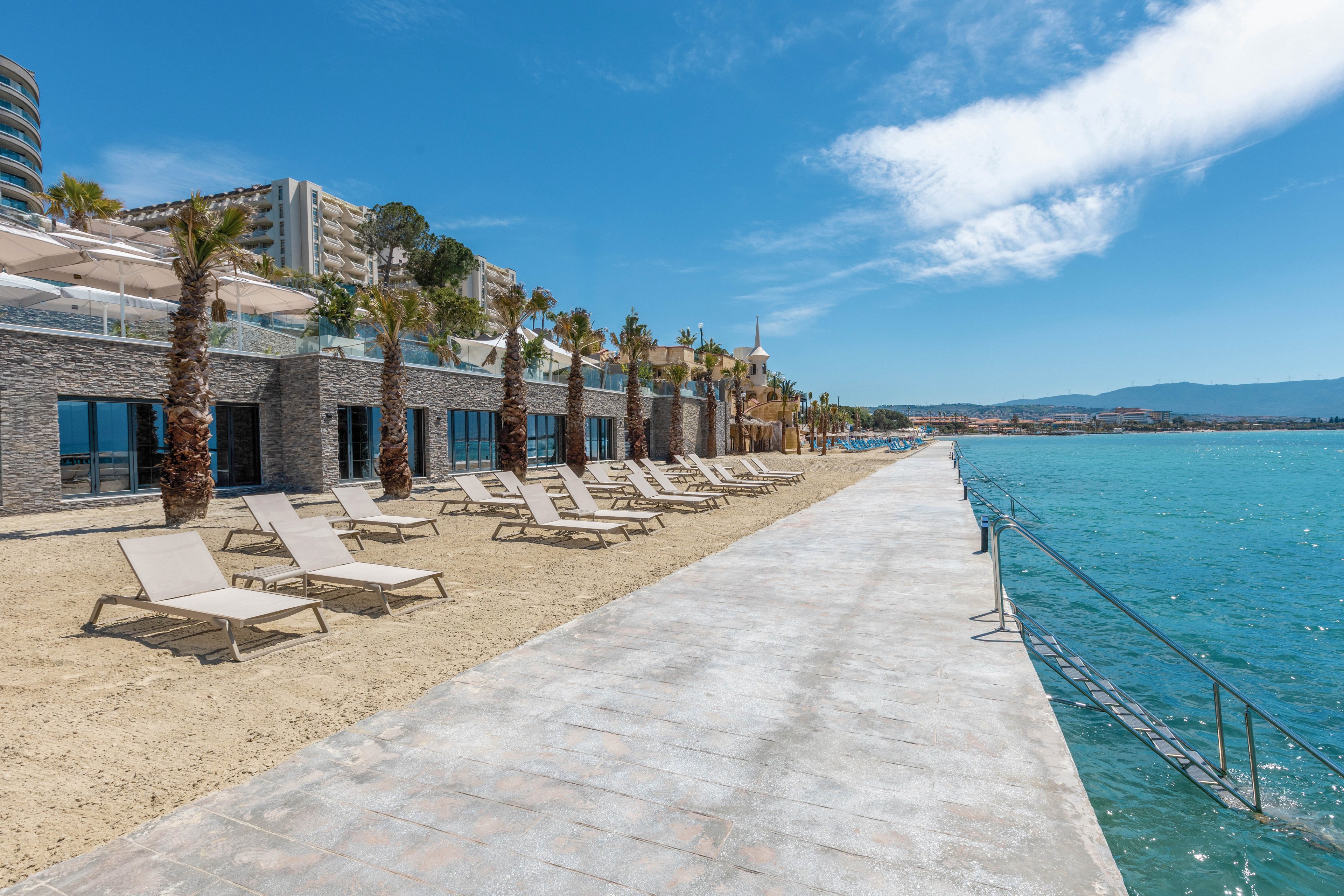 A modern seaside resort features rows of empty sun loungers arranged on a sandy strip beside a wide concrete promenade. Palm trees line the waterfront, with contemporary buildings and terraces rising behind them. Calm turquoise water borders the walkway, with metal steps leading directly into the sea. The bright blue sky and scattered clouds create a sunny, tranquil vacation atmosphere.