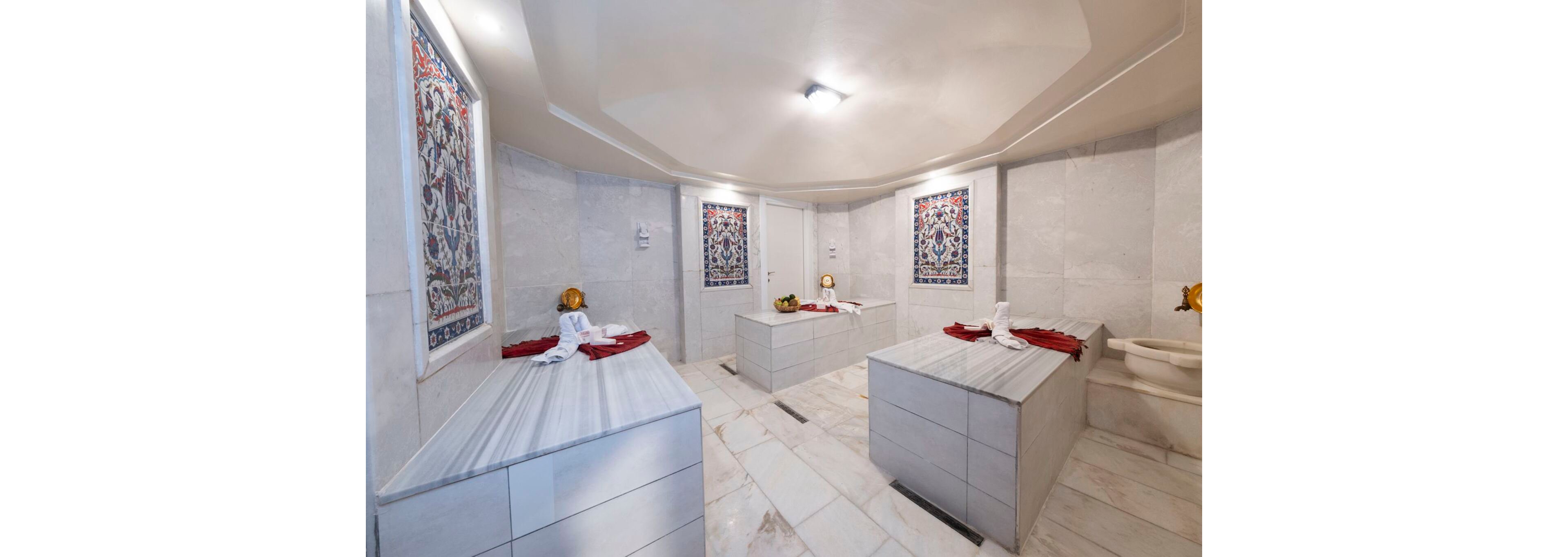 Bright marble-clad Turkish bath room with multiple massage platforms dominates the scene. Ornate tiled wall panels add traditional decorative detail to the clean white surfaces. Rolled white towels and red cloths are neatly arranged on the stone slabs, suggesting spa service readiness. Soft ceiling lighting creates a calm, relaxing atmosphere in the spacious hammam.