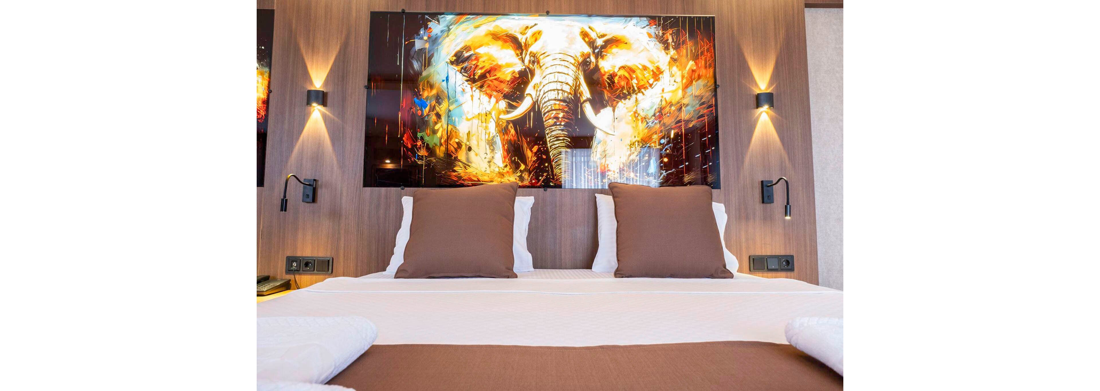 A neatly made double bed with white linens and brown pillows is positioned against a wooden accent wall. Above the headboard hangs a large, colorful painting of an elephant with fiery abstract effects. Wall-mounted reading lights flank the artwork, adding a warm, contemporary feel to the room. The overall setting suggests a stylish, modern hotel or home interior focused on comfort and bold decor.