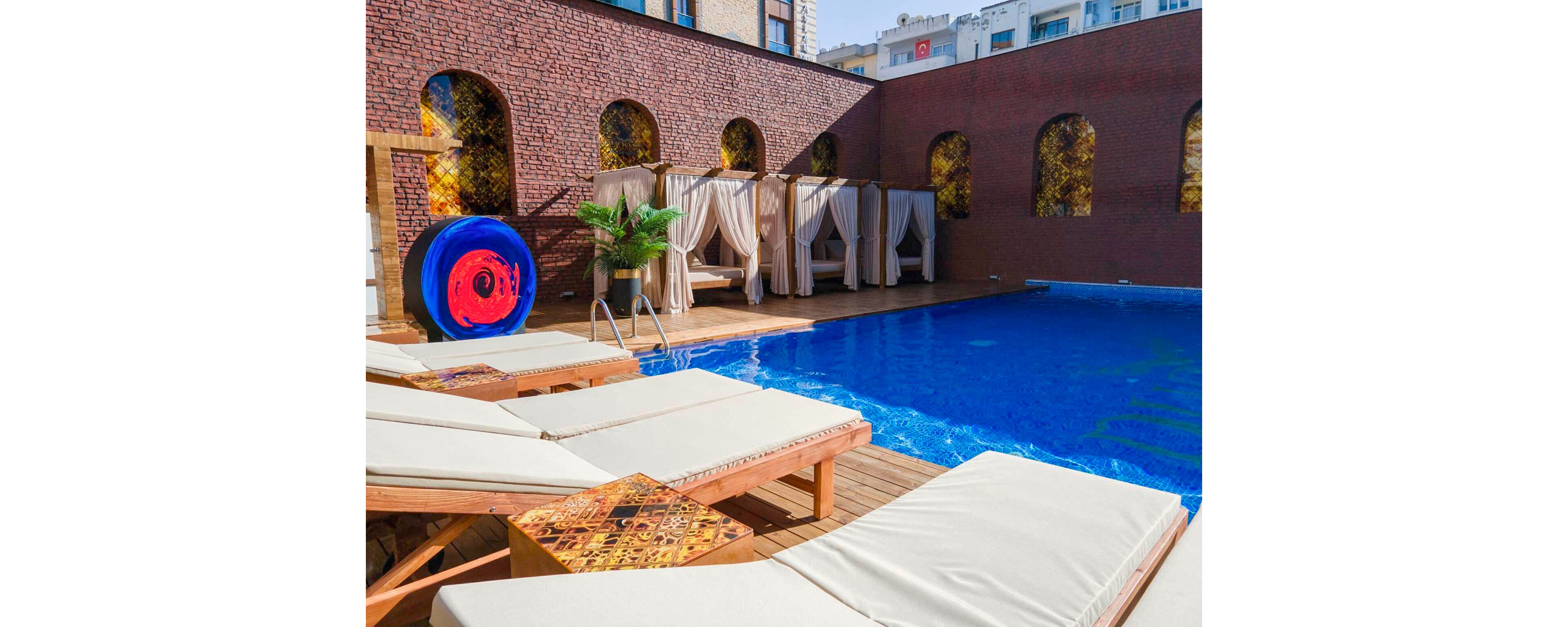 Outdoor swimming pool area with cushioned wooden loungers and a bright blue circular art piece near the ladder. Elegant cabanas with flowing curtains line the brick wall, creating a private retreat atmosphere. The vivid blue water contrasts with warm wood decking and textured brick architecture. City buildings are visible in the background, suggesting a rooftop or courtyard setting ideal for relaxation.