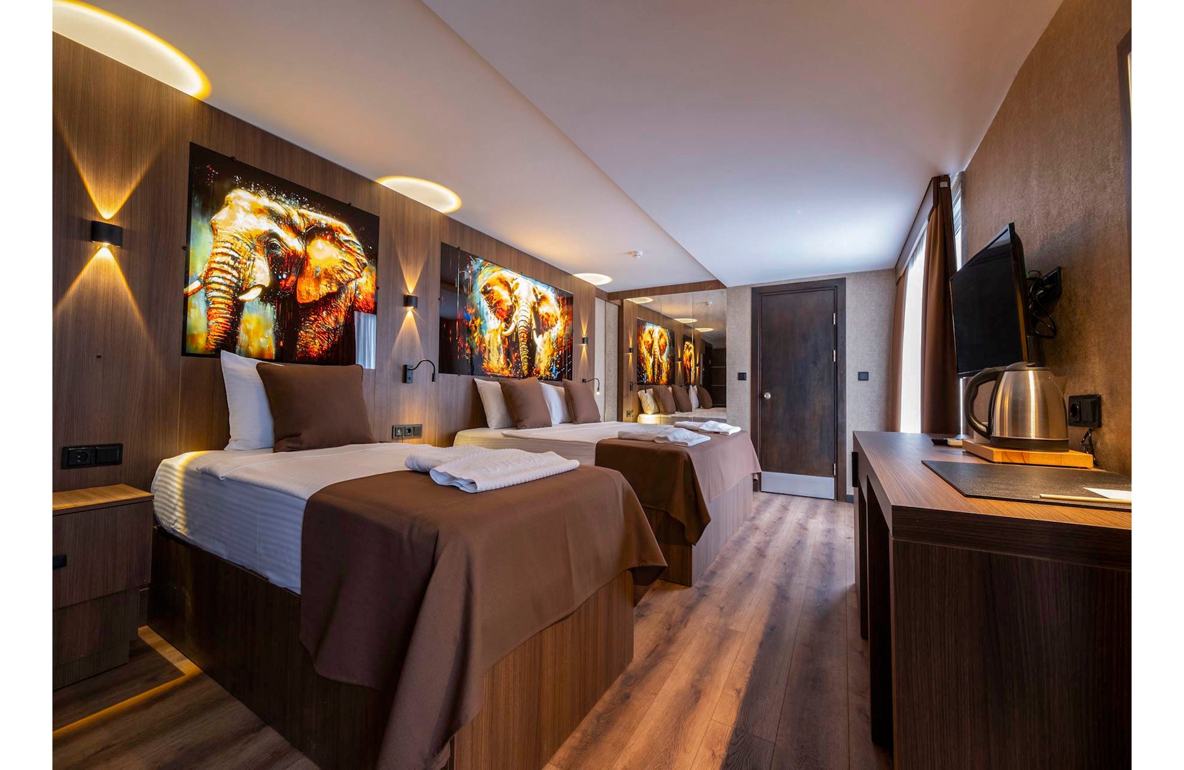 A contemporary hotel room features multiple beds neatly arranged with brown bedspreads and white linens. Vibrant illuminated wall art depicting elephants adds a dramatic focal point above the beds. Warm lighting and wood finishes create a cozy, inviting atmosphere, complemented by a flat-screen TV and electric kettle on the desk. The room extends toward a doorway at the far end, emphasizing its spacious layout.