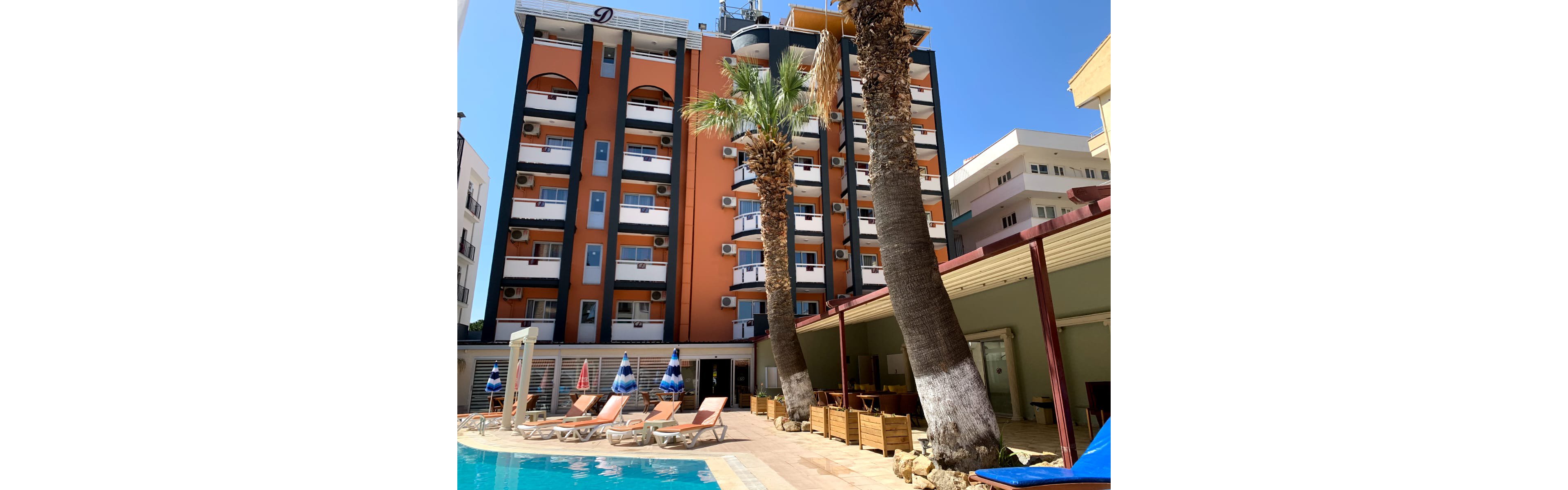 A multi-story orange and white hotel rises behind a small outdoor swimming pool. Sun loungers and blue-and-white umbrellas line the poolside terrace, shaded partly by tall palm trees. A covered seating area with wooden furniture runs along one side, while neighboring apartment buildings frame the scene. Bright daylight and clear blue sky create a warm, inviting vacation atmosphere.