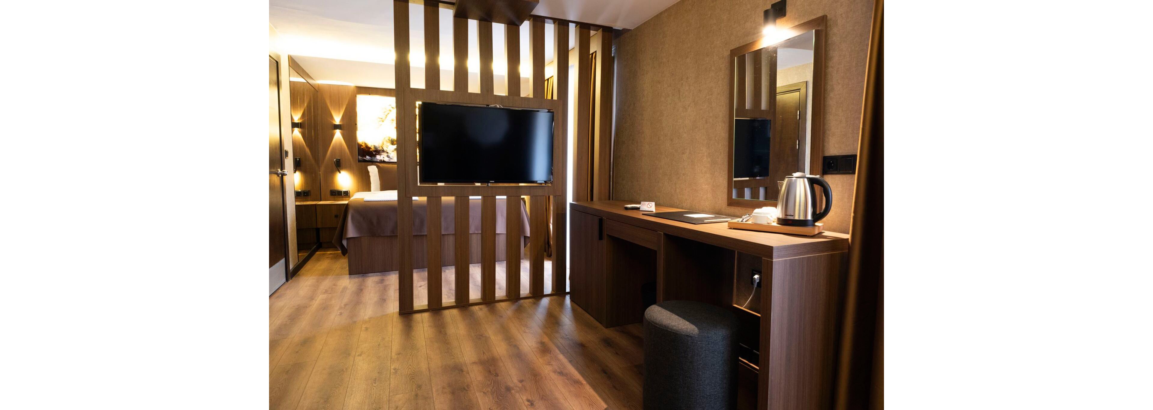 Contemporary hotel room features a wooden partition with a flat-screen television separating the sleeping area from the workspace. A long wooden desk with a cushioned stool sits beside the partition, illuminated by a wall-mounted light. A metal electric kettle and cups are arranged neatly on the desk, suggesting comfort and hospitality. Warm brown tones, soft lighting, and wood flooring create a cozy, modern atmosphere.