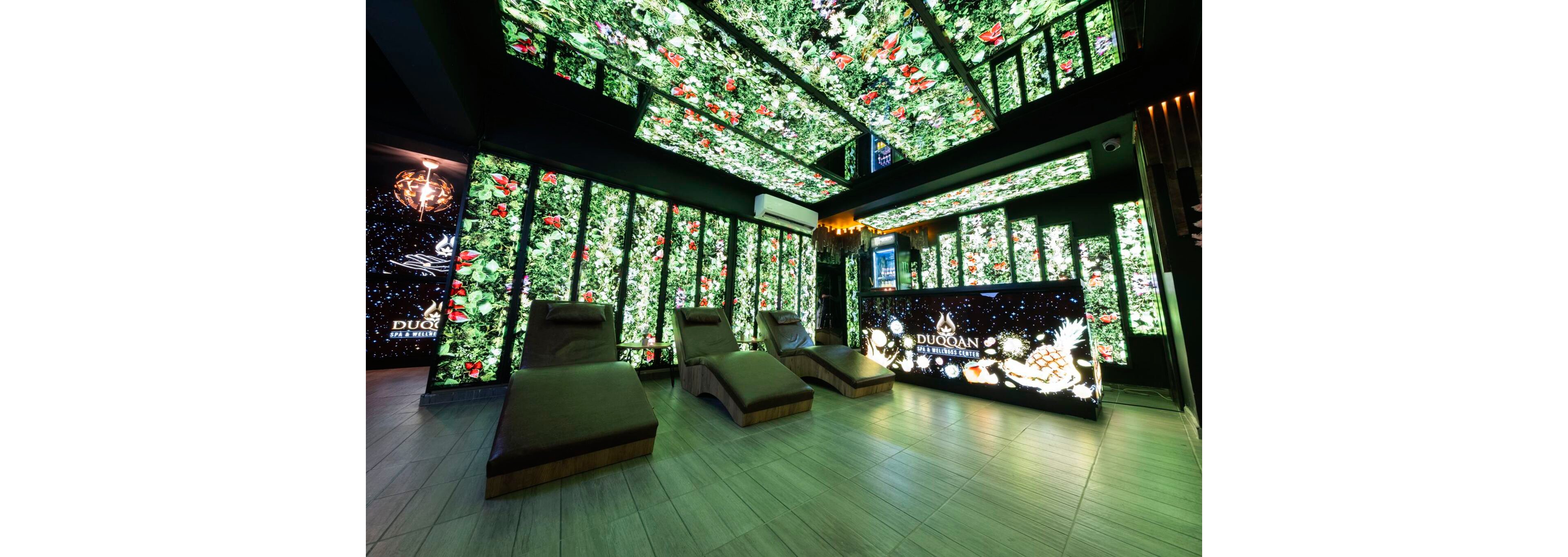A contemporary spa lounge features reclining chaise chairs arranged on a light wood floor. The walls and ceiling are covered with bright illuminated panels displaying lush green foliage and red flowers, creating an immersive garden effect. At the back of the room, a reception counter with glowing fruit graphics and the word "DUOCAN" stands out against the vibrant backdrop. The overall atmosphere is luxurious, futuristic, and relaxing.