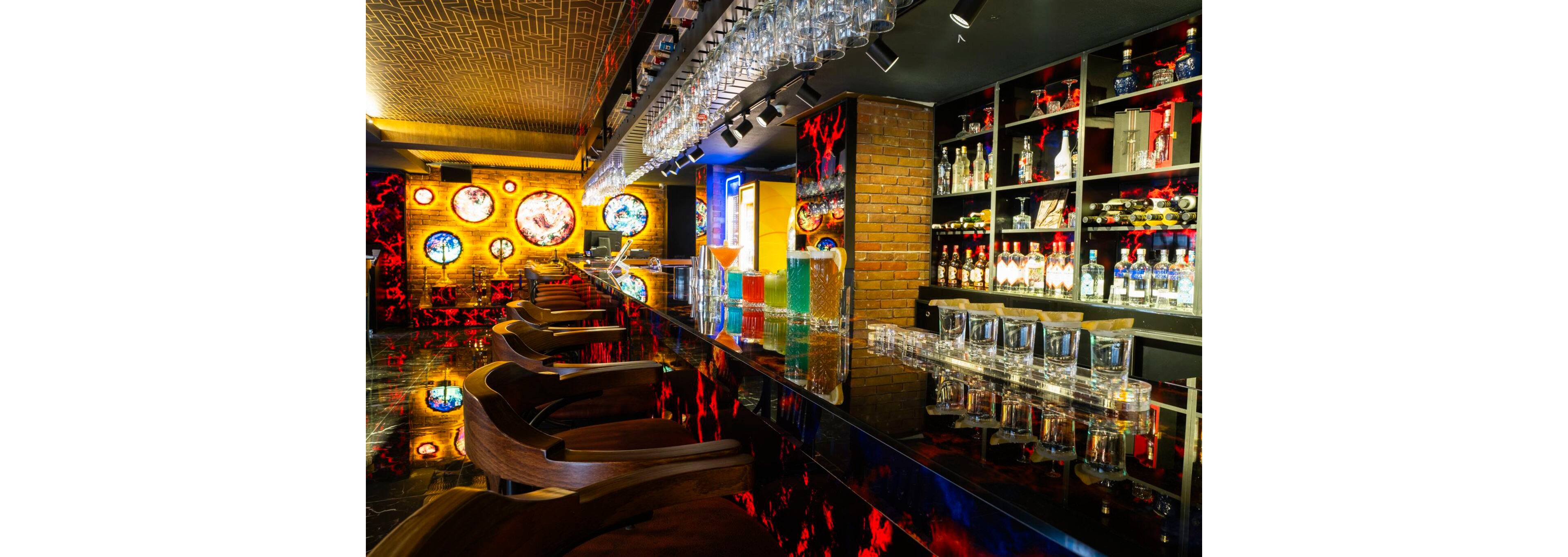 A modern bar interior features a long glossy counter lined with wooden bar stools and rows of empty glasses. Colorful cocktails in tall glasses stand on the bar, reflecting vibrant hues on the shiny surface. The background showcases shelves filled with liquor bottles and brick walls adorned with circular illuminated artworks. Warm lighting and neon accents create an energetic, nightlife atmosphere.
