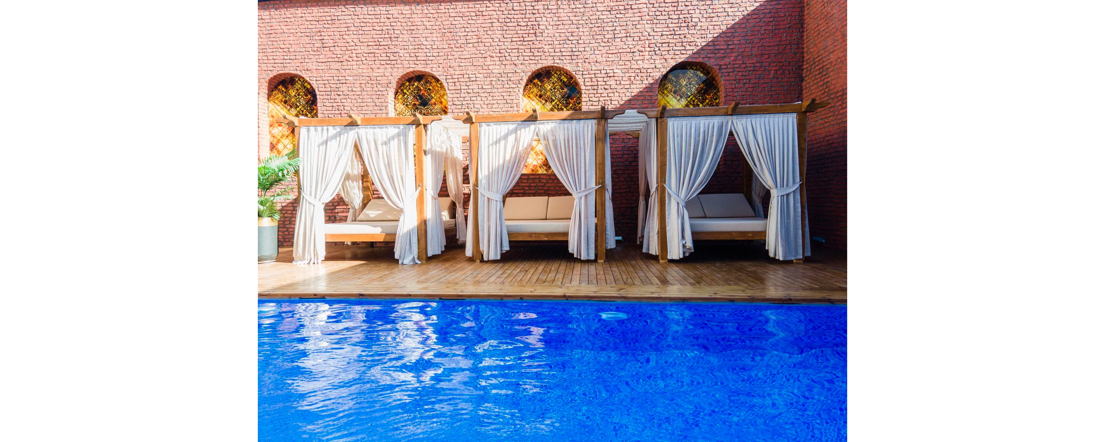The image shows a row of elegant poolside cabanas with flowing white curtains and cushioned lounge beds. They are set against a red brick wall featuring arched windows with decorative stained glass. A bright blue swimming pool occupies the foreground, reflecting the vivid color of the water. The overall scene suggests a relaxing, upscale resort environment with warm, inviting tones.