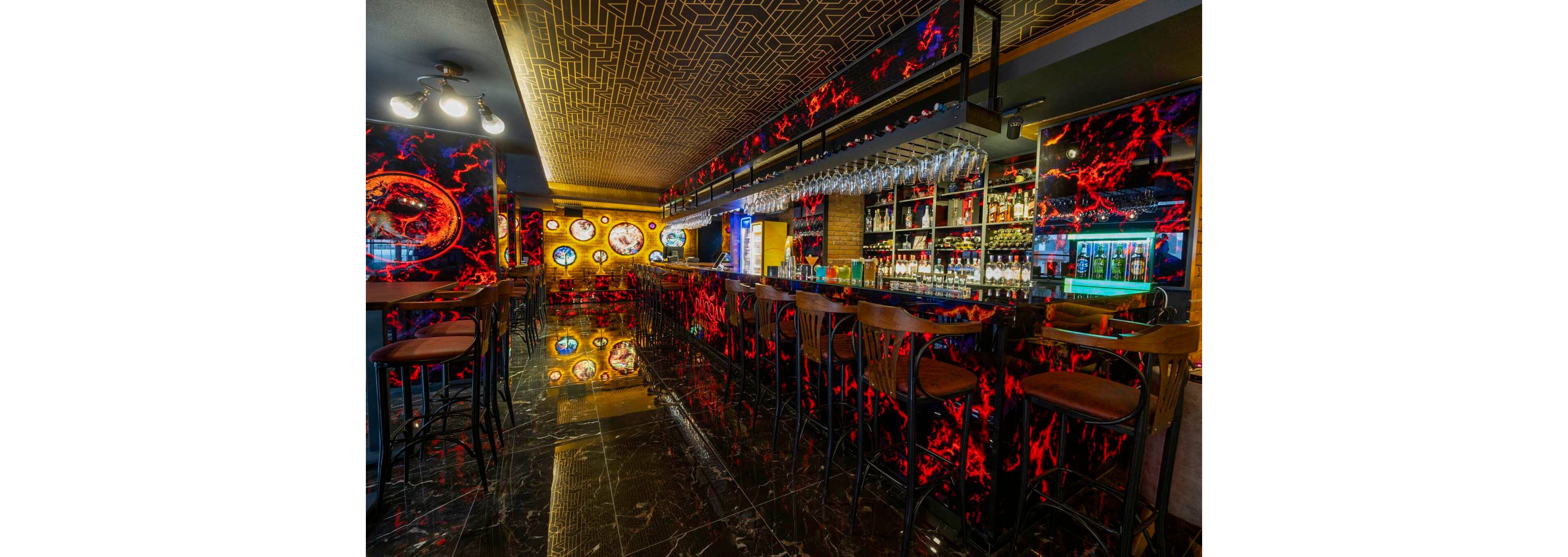 A stylish cocktail bar interior features a long counter with high wooden stools and dramatic neon-red abstract wall panels. Shelves behind the bar are filled with assorted bottles, glasses, and backlit displays, creating a vibrant nightlife atmosphere. The ceiling has a geometric gold pattern, while the glossy dark floor reflects the colorful lights and decor. Circular illuminated art pieces and hanging glassware add a modern, immersive feel to the space.