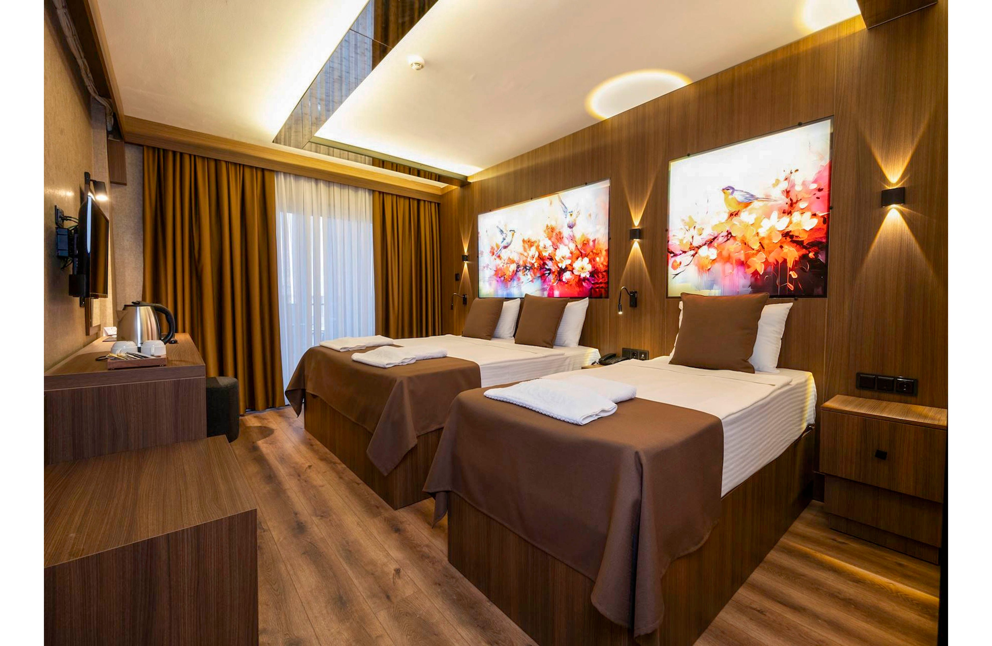 Contemporary hotel guest room featuring two neatly made twin beds with brown bedspreads and white linens. The space is accented by warm wooden paneling, floor-to-ceiling curtains, and soft ambient lighting. Colorful wall art with birds and floral motifs adds a vibrant focal point above the beds. A small desk area with a kettle and mounted television completes the cozy, well-appointed interior.