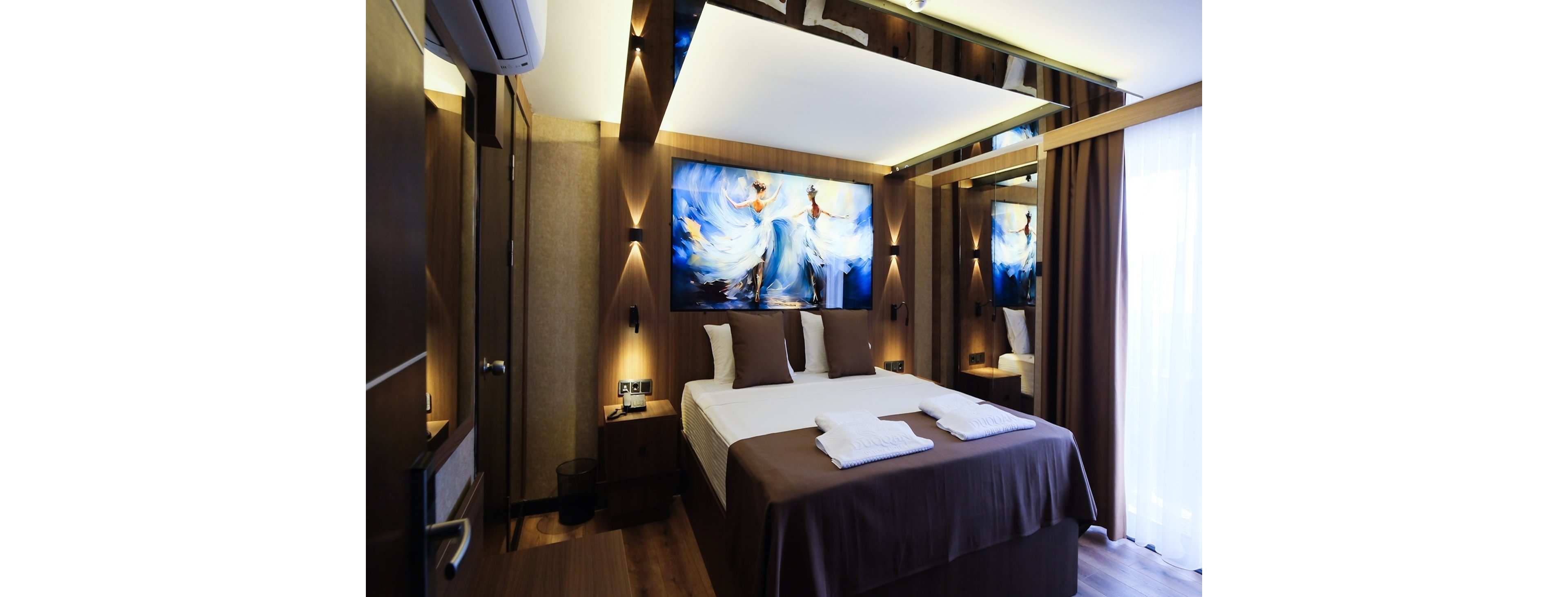 Contemporary double hotel room featuring a neatly made bed with brown linens and white pillows. The space is accented by a large colorful painting of dancing figures above the headboard. Warm recessed lighting, wood paneling, and floor-to-ceiling curtains create a cozy, upscale atmosphere. A bedside table, wall-mounted reading lights, and folded white towels on the bed suggest comfort and readiness for guests.