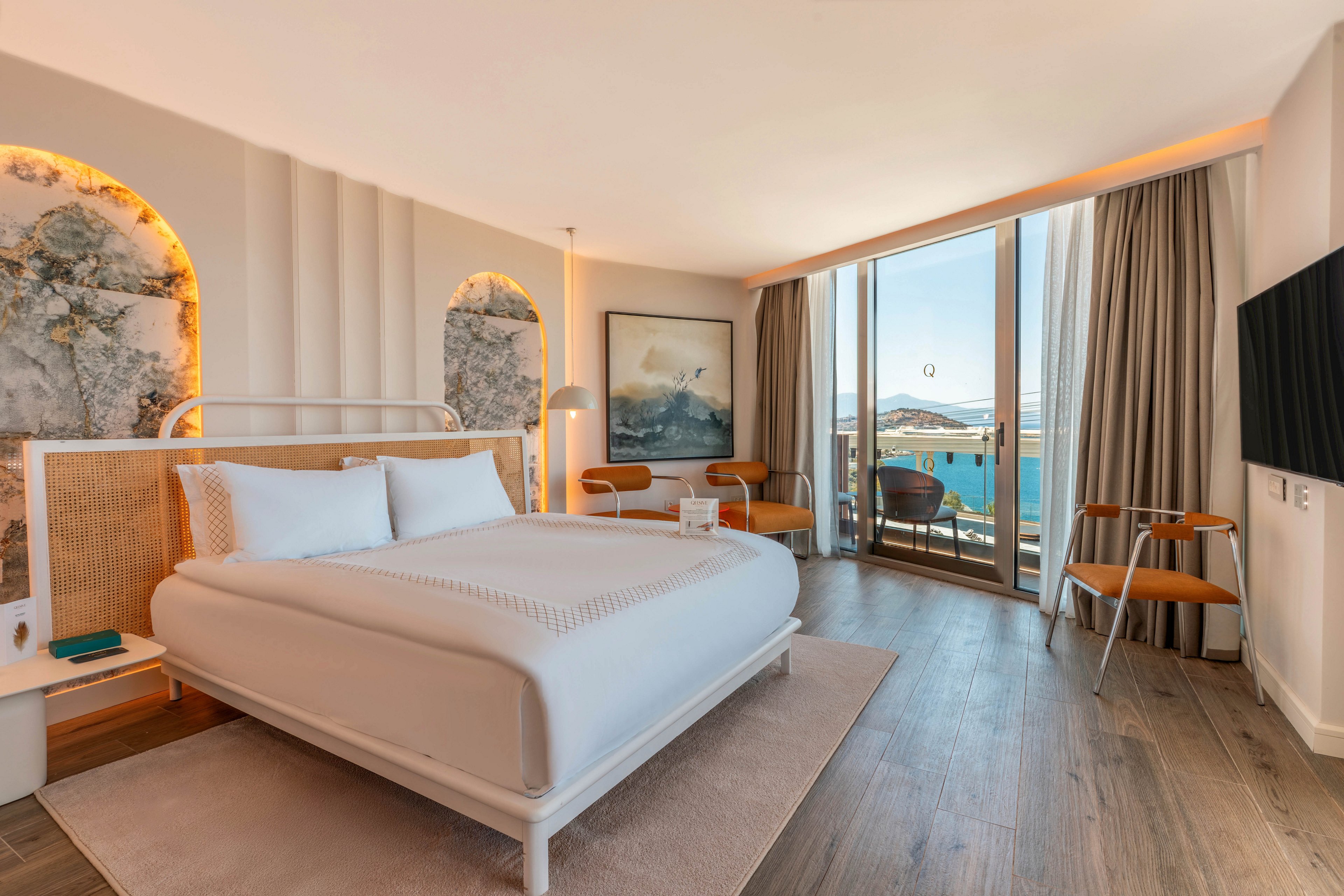 A contemporary hotel bedroom features a neatly made double bed with white linens and a light wood headboard. The room includes minimalist furniture such as a side table, chairs, and wall art, all arranged on a wooden floor. Large floor-to-ceiling windows open onto a balcony overlooking a bright blue ocean and distant hills. Warm ambient lighting and neutral tones create a calm, inviting atmosphere.