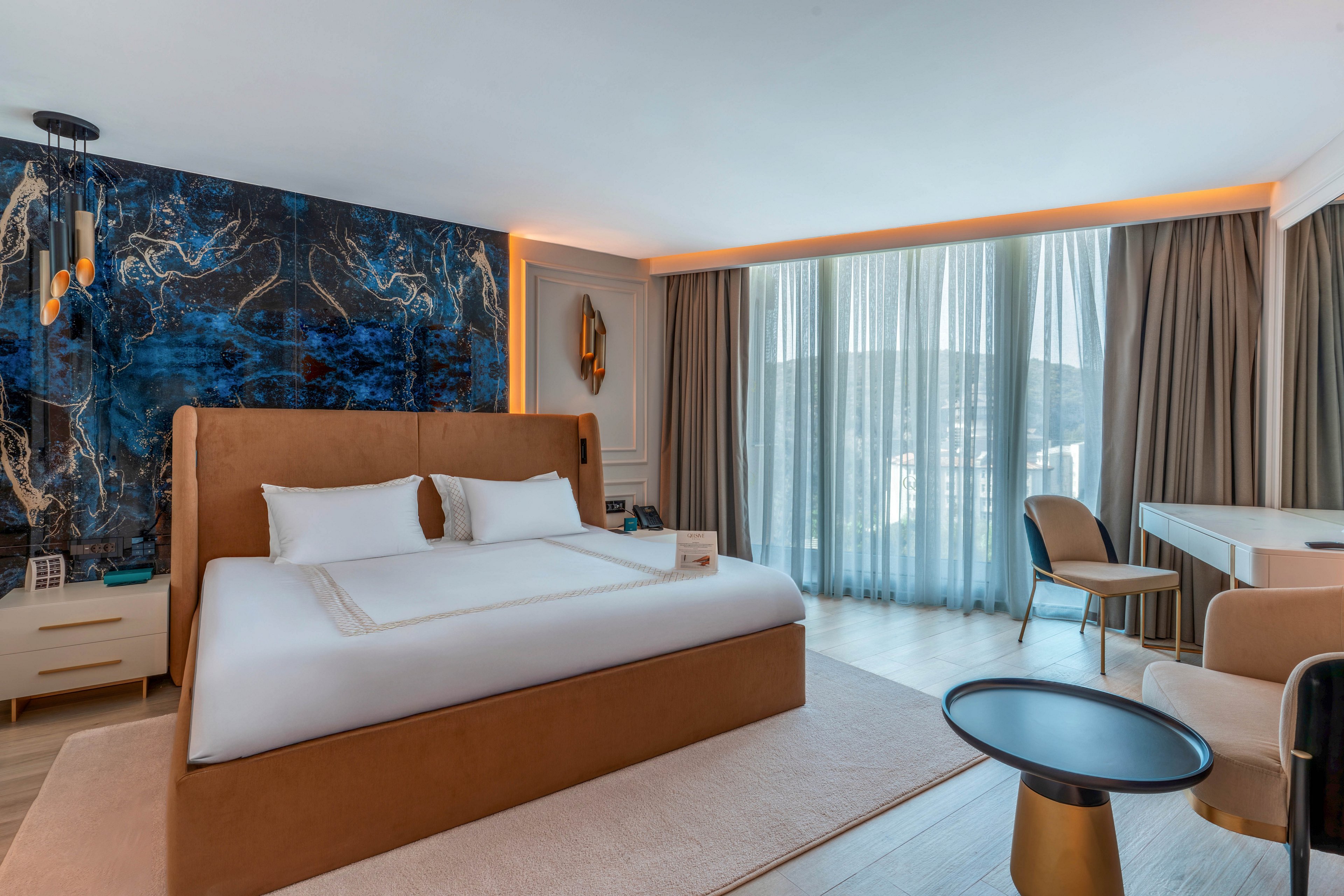 A contemporary hotel bedroom features a large bed with a tan upholstered frame and crisp white linens. The room includes a striking blue abstract accent wall behind the headboard and warm ambient lighting. Floor-to-ceiling sheer curtains cover a wide window, allowing soft daylight to filter into the space. A small seating area with modern chairs and tables, along with a sleek desk, completes the stylish and comfortable interior.
