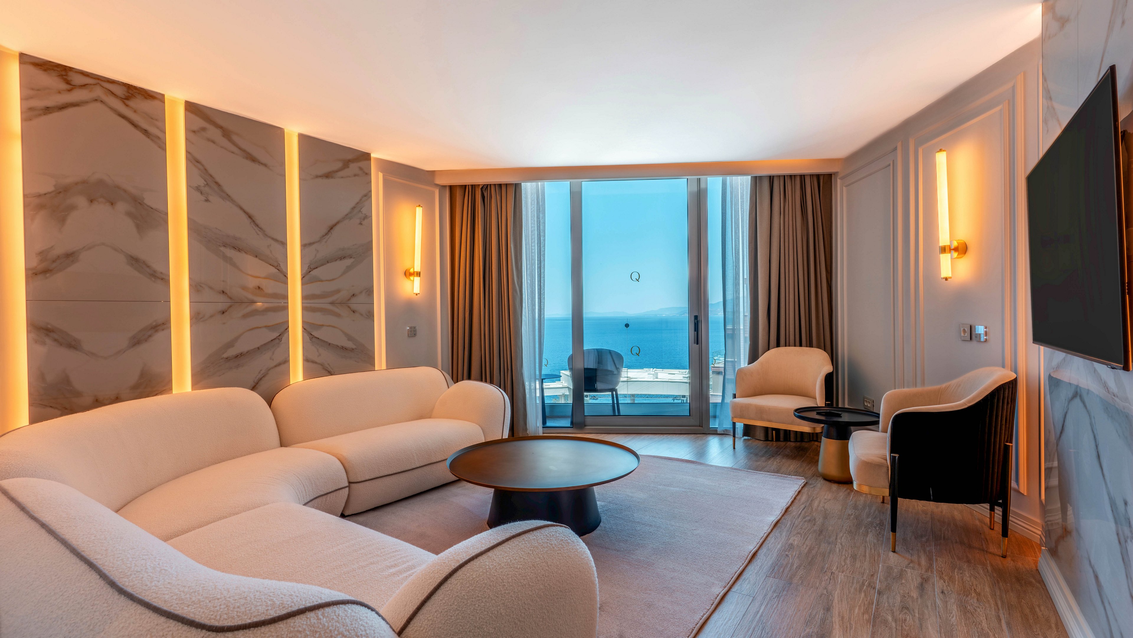 A contemporary hotel suite features curved cream sofas arranged around a round coffee table. Warm vertical light strips accent marble wall panels, creating a sophisticated ambiance. Floor-to-ceiling windows and glass doors open to a balcony overlooking a bright blue ocean. Neutral tones, soft upholstery, and wooden flooring contribute to a calm, upscale interior setting.