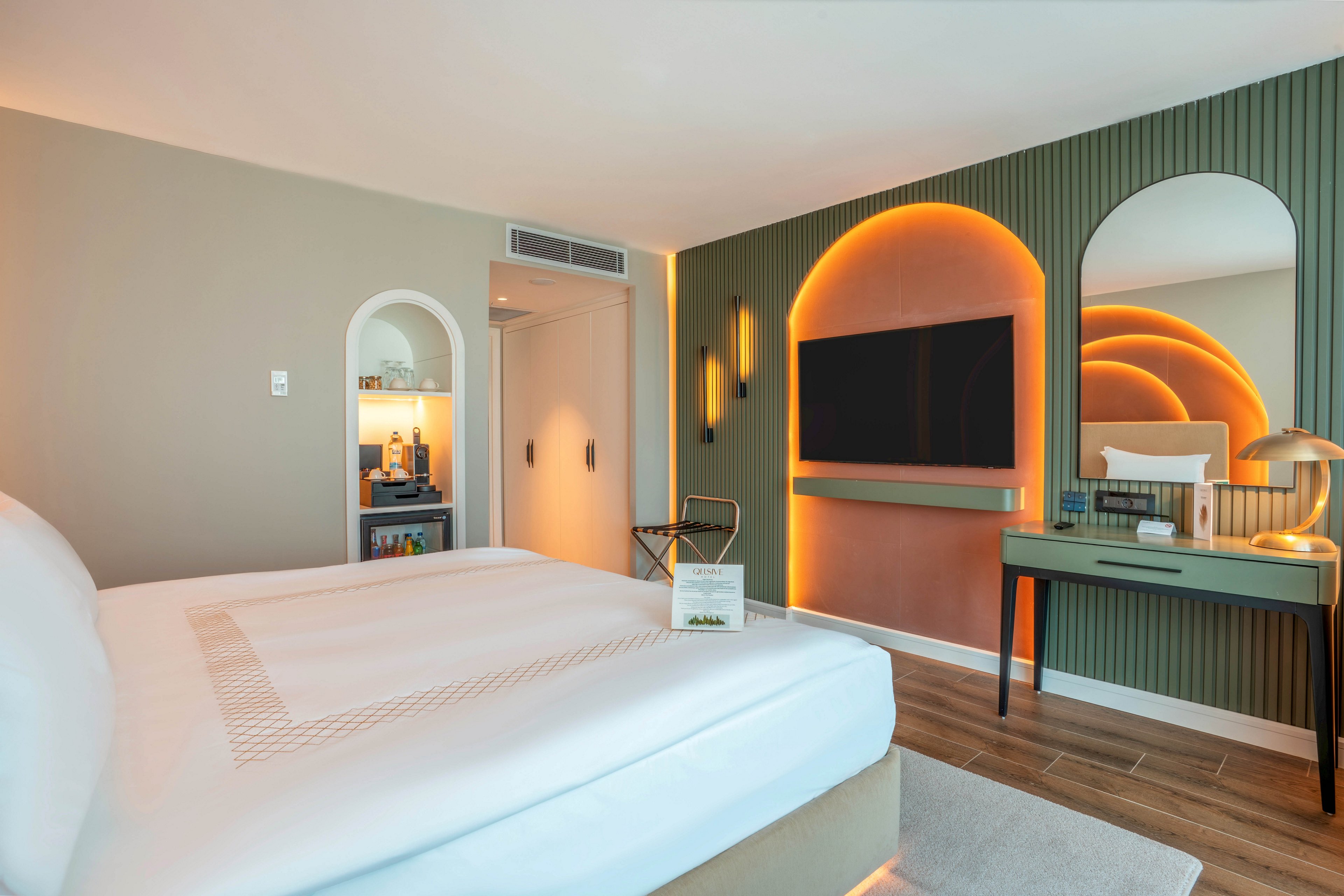 A contemporary hotel room features a neatly made bed in the foreground facing a wall-mounted flat-screen television. The accent wall behind the TV has warm orange backlighting in arched shapes, creating a stylish focal point. To the right, a desk with a mirror and decorative lamp adds functionality, while a small open shelving nook with beverages and glasses sits near the door. Soft neutral tones, wood flooring, and subtle lighting contribute to a cozy yet modern atmosphere.