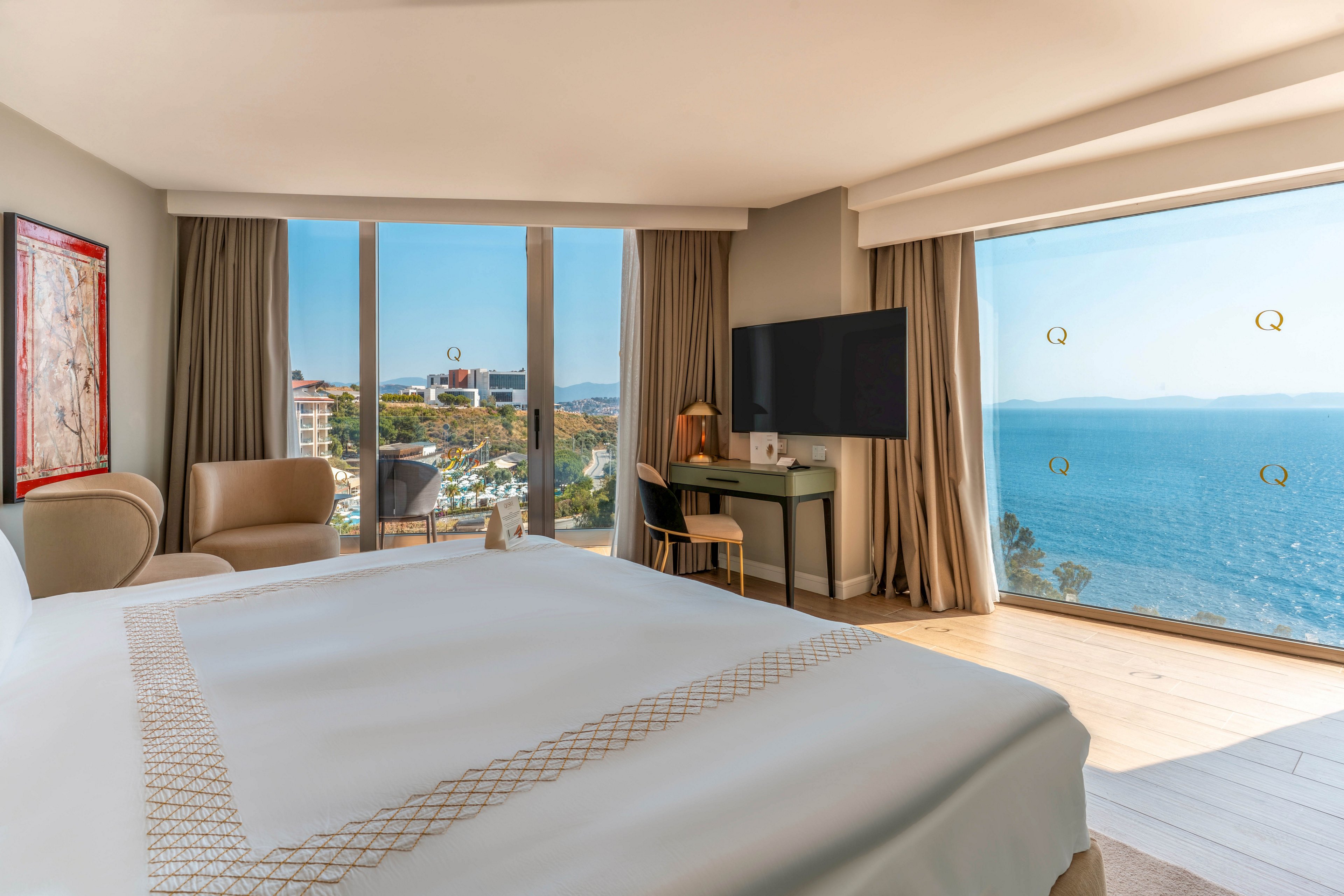 A contemporary hotel bedroom features a large neatly made bed facing floor-to-ceiling windows. The room includes a small seating area with two chairs and a desk with a television mounted above it. Beyond the windows, a coastal town and expansive blue sea are visible under clear skies. Neutral tones and natural light create a calm, airy atmosphere ideal for relaxation.