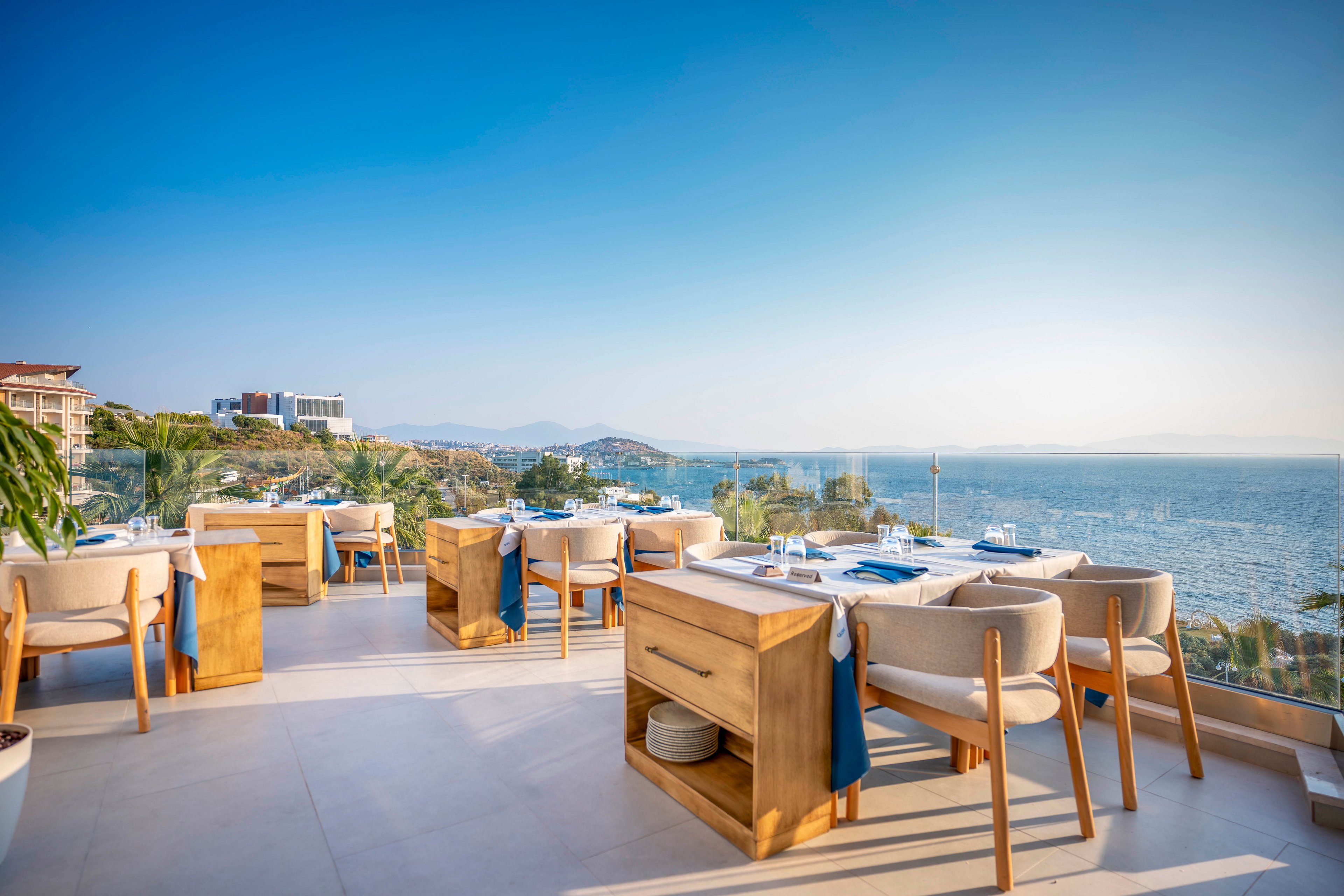 An elegant open-air restaurant terrace overlooks a calm blue sea. Light wooden tables and cushioned chairs are neatly arranged along a glass railing, creating a modern and inviting dining space. The clear sky and bright sunlight enhance the coastal scenery, with boats and distant buildings visible along the shoreline. The overall atmosphere is relaxed, luxurious, and ideal for al fresco dining by the water.