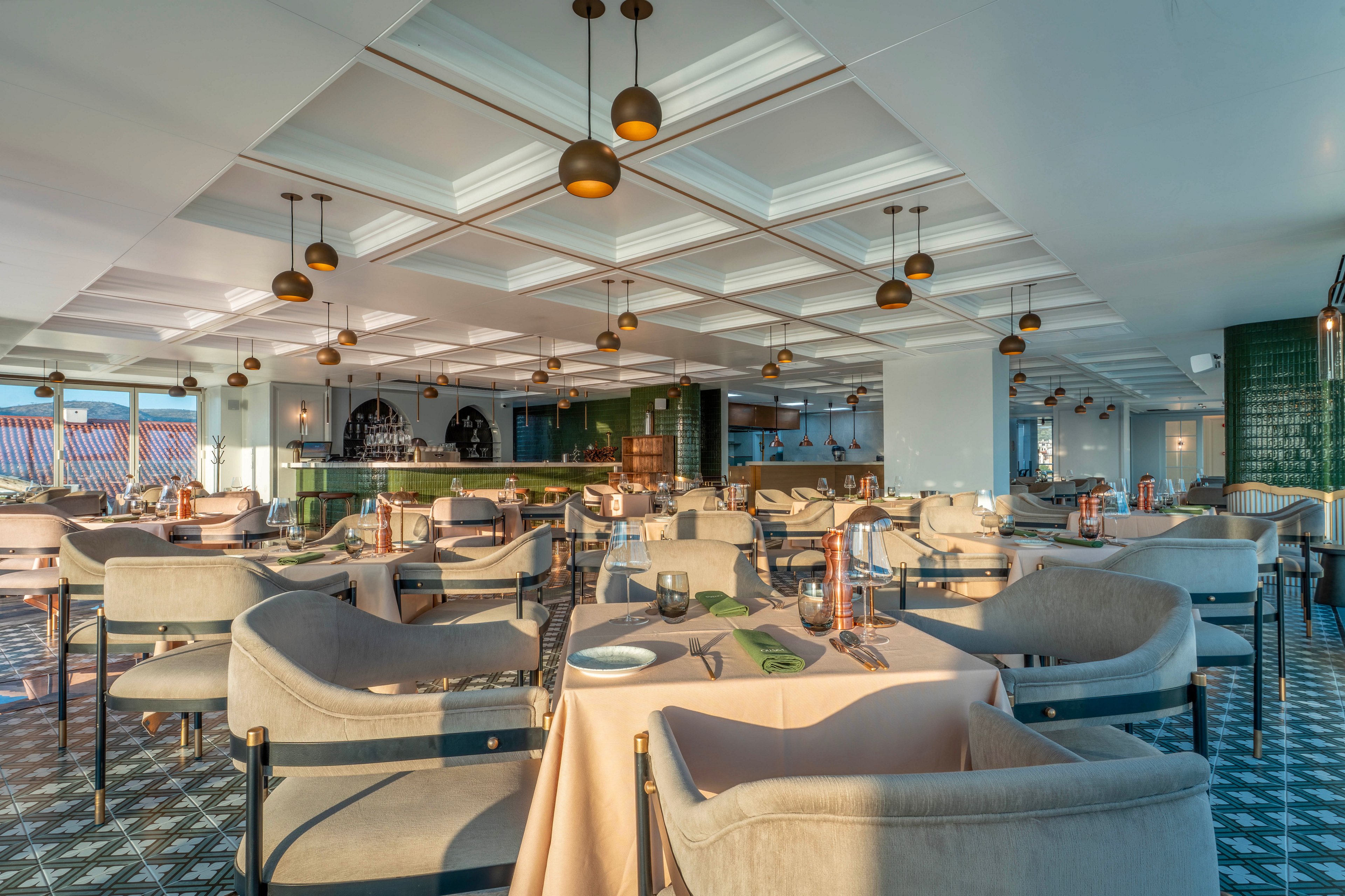 A spacious contemporary restaurant interior features neatly arranged tables and upholstered chairs in soft neutral tones. Large windows allow abundant natural light to flood the room, highlighting the patterned floor tiles and coffered ceiling. The open bar area with green accents and warm pendant lighting creates a stylish focal point at the back. The overall atmosphere feels calm, inviting, and ready for guests.