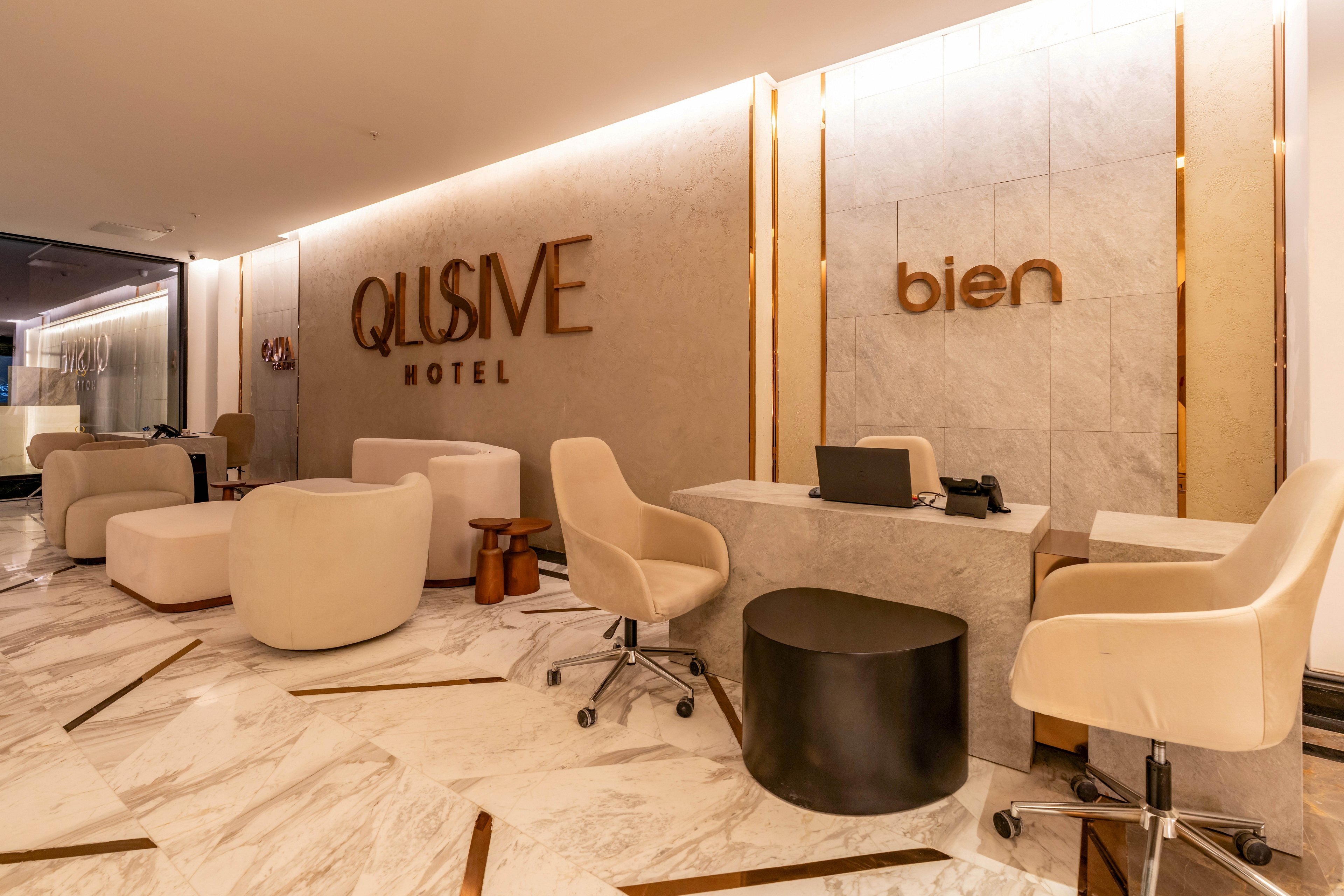 Contemporary hotel lobby with a sleek reception desk and plush seating is shown. The space features beige armchairs, rounded lounge chairs, and a black cylindrical side table on a polished marble floor. Warm ambient lighting highlights the wall with the words "QLUSIVE HOTEL" and "bien" in metallic letters. The overall design feels upscale, minimalist, and welcoming for arriving guests.