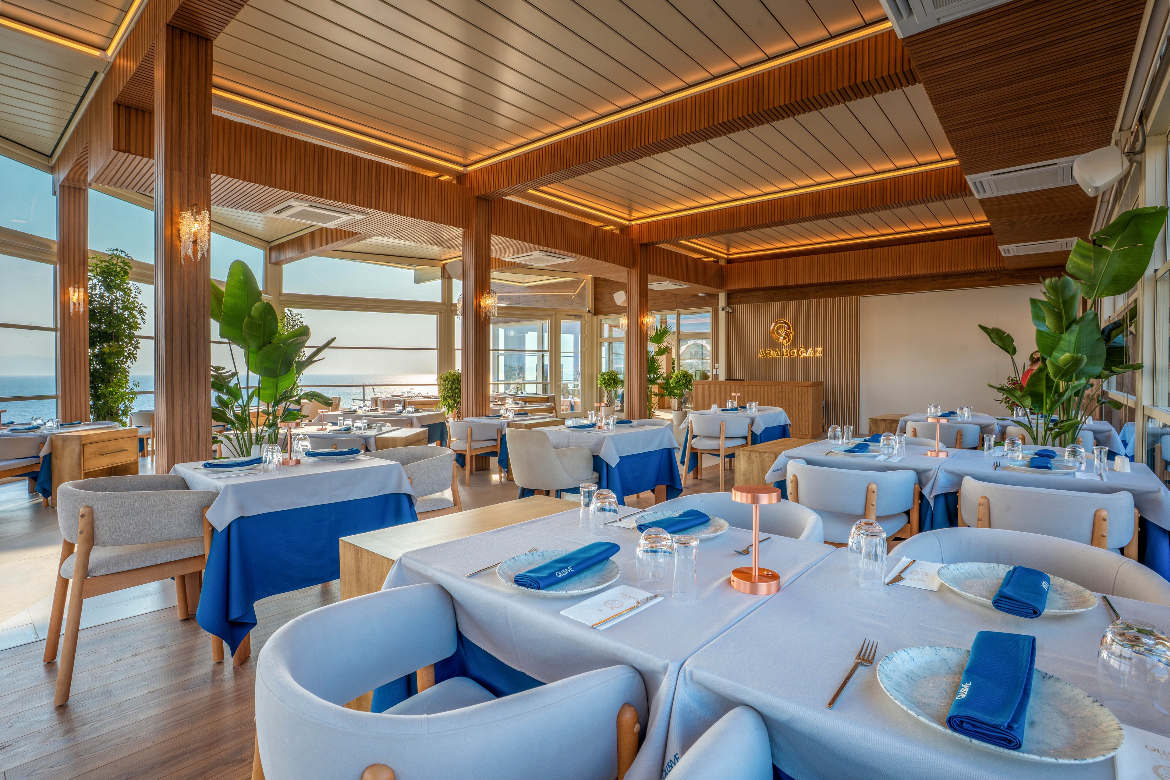 A contemporary restaurant dining room is set with neatly arranged tables covered in white cloths and blue table runners. Soft upholstered chairs surround the tables, each laid with plates, glasses, napkins, and cutlery ready for guests. Large windows and open doors reveal a bright seaside view, filling the space with natural light. Warm ceiling lighting, wooden accents, and lush green plants create a relaxed yet upscale coastal atmosphere.