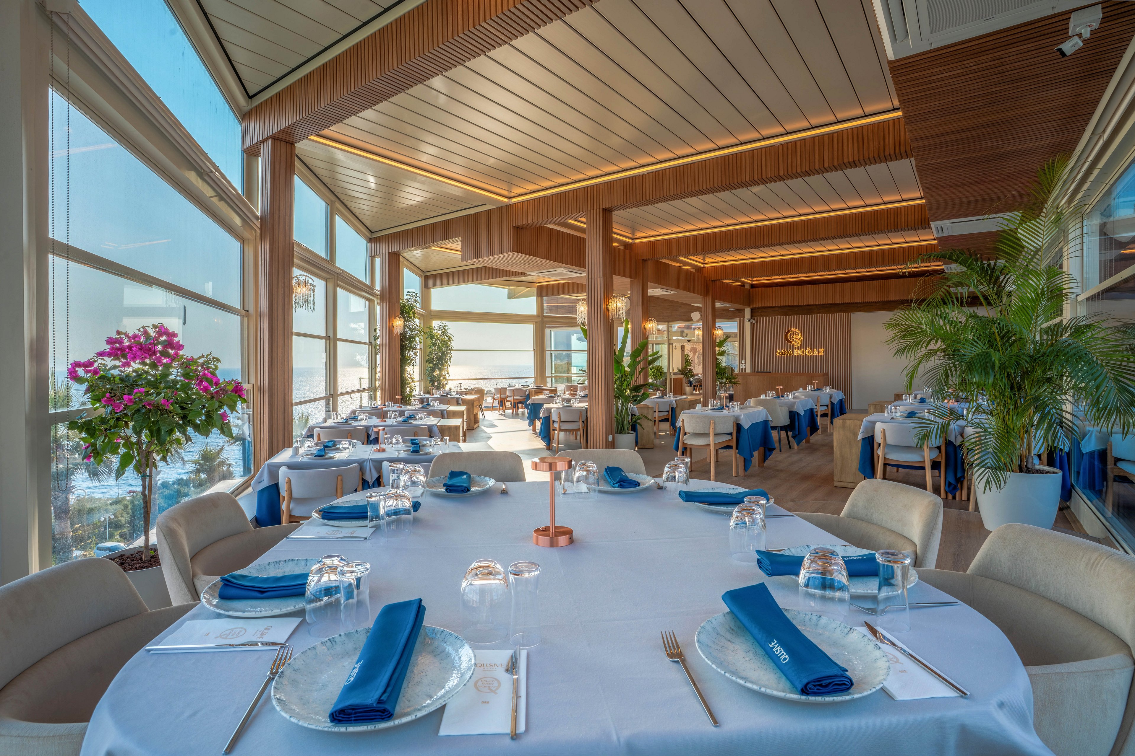 A bright, upscale restaurant dining room is set with round tables covered in white linens and neatly folded blue napkins. Large floor-to-ceiling windows reveal a coastal view, filling the space with natural light. Warm wooden ceilings, modern lighting, and lush green plants create a sophisticated yet relaxed atmosphere. The room is empty of guests, emphasizing the orderly table settings and serene seaside ambiance.