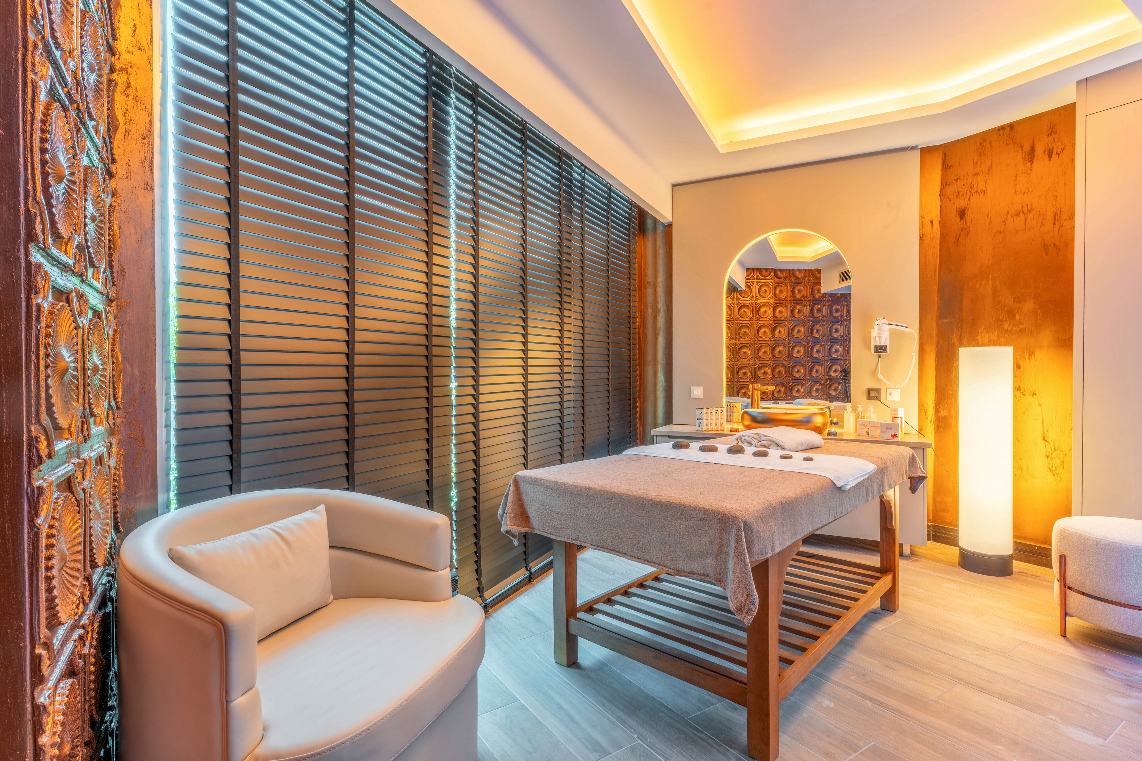 A serene spa treatment room features a massage table neatly prepared with folded towels. The space is illuminated by warm ambient lighting from the ceiling and a tall floor lamp, creating a relaxing atmosphere. Decorative wall panels and an arched mirror add an elegant touch to the interior design. A comfortable armchair and wooden blinds complete the cozy, modern wellness setting.