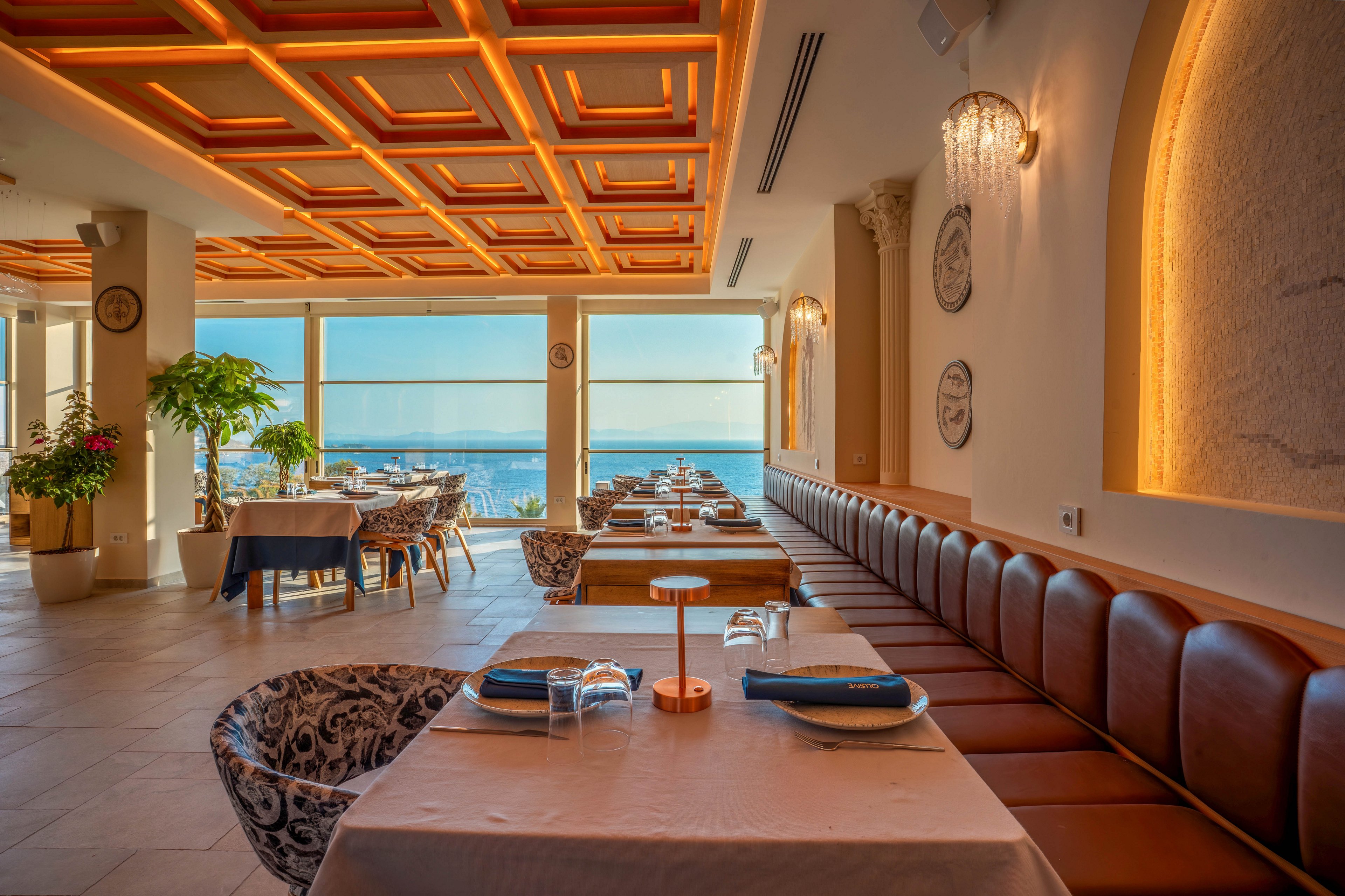 A stylish restaurant interior features neatly arranged tables with folded napkins and glassware. Large floor-to-ceiling windows reveal a scenic view of the sea and a coastal city beyond. Warm ceiling lights and wall sconces create a cozy, golden ambiance that contrasts with the cool blue tones outside. Leather banquette seating and patterned chairs add a touch of luxury to the contemporary dining space.
