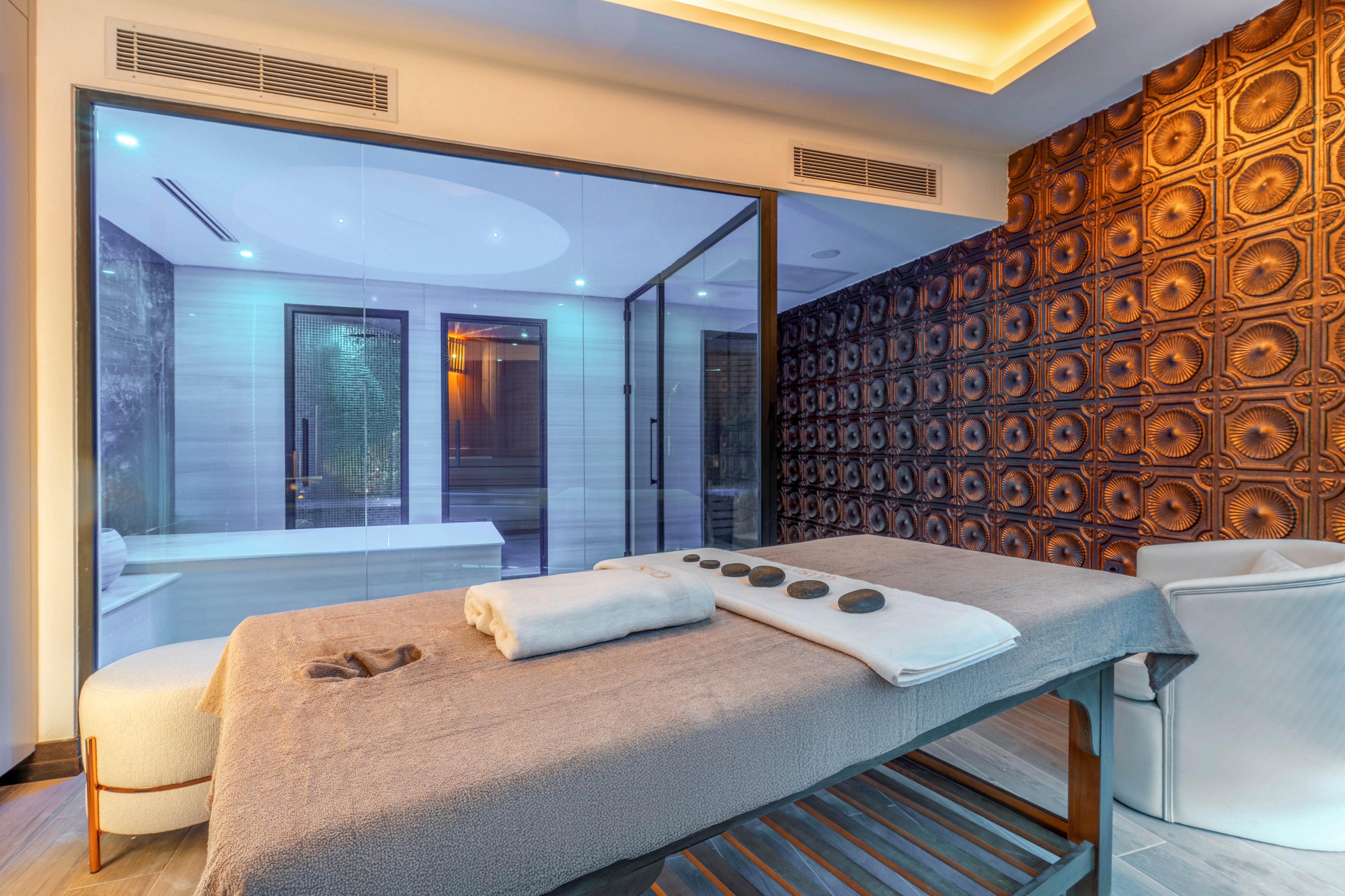 A contemporary spa treatment room features a massage table prepared with folded towels and hot stones. The space is enclosed by a large glass wall that reveals a sleek adjoining bathroom with soft ambient lighting. Warm wooden accents and a textured decorative wall create a sophisticated, upscale atmosphere. Cool blue and warm yellow lights combine to enhance the relaxing, tranquil mood of the interior.