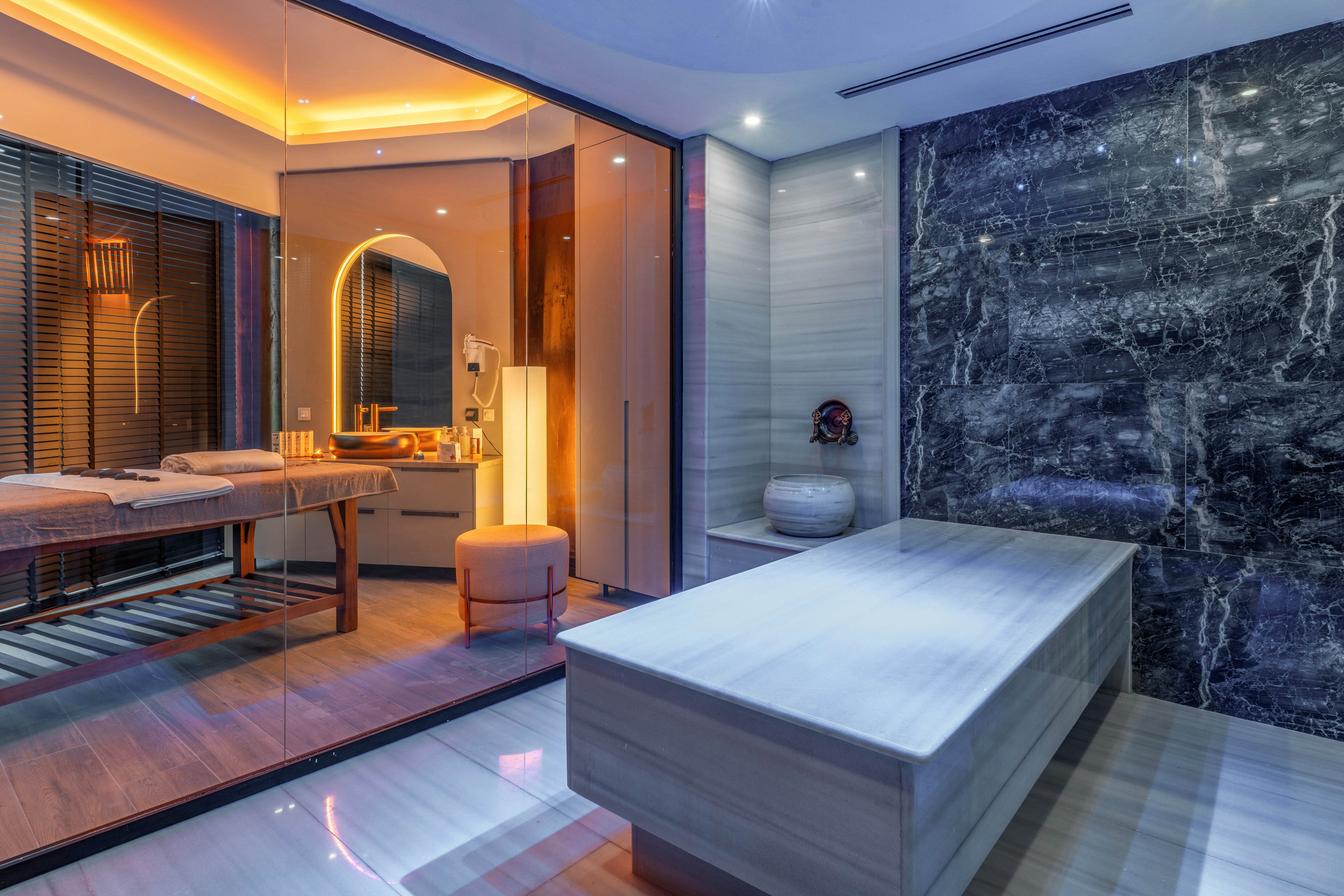 A contemporary spa interior features a sleek marble treatment table in the foreground. Behind a glass partition, a warm-lit massage room with a wooden table and stool creates a cozy contrast. Dark stone walls and reflective surfaces add a sophisticated, high-end feel to the space. The combination of cool marble and ambient lighting emphasizes relaxation and wellness.