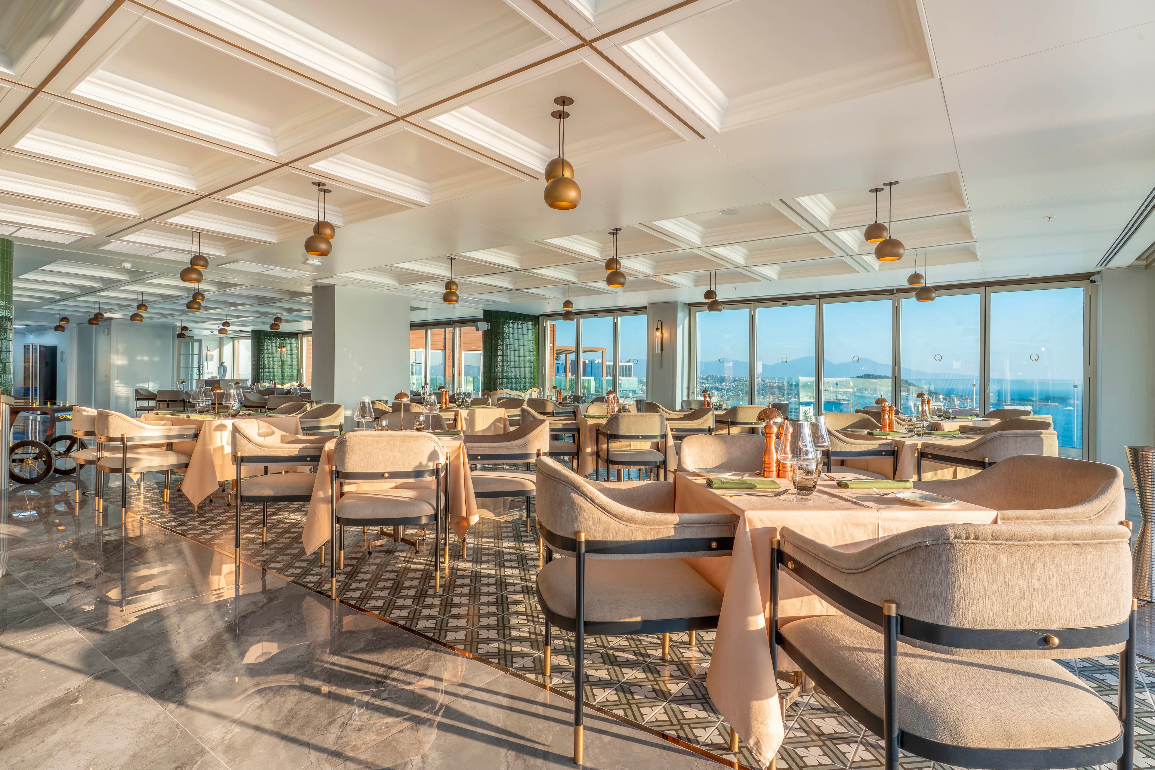 A spacious modern restaurant dining area is filled with neatly arranged tables and upholstered chairs. Large floor-to-ceiling windows line the wall, offering expansive views of the city and distant mountains. Warm sunlight floods the room, highlighting the neutral tones, metallic accents, and polished stone floor. The setting appears calm and inviting, ready to welcome guests for a meal.