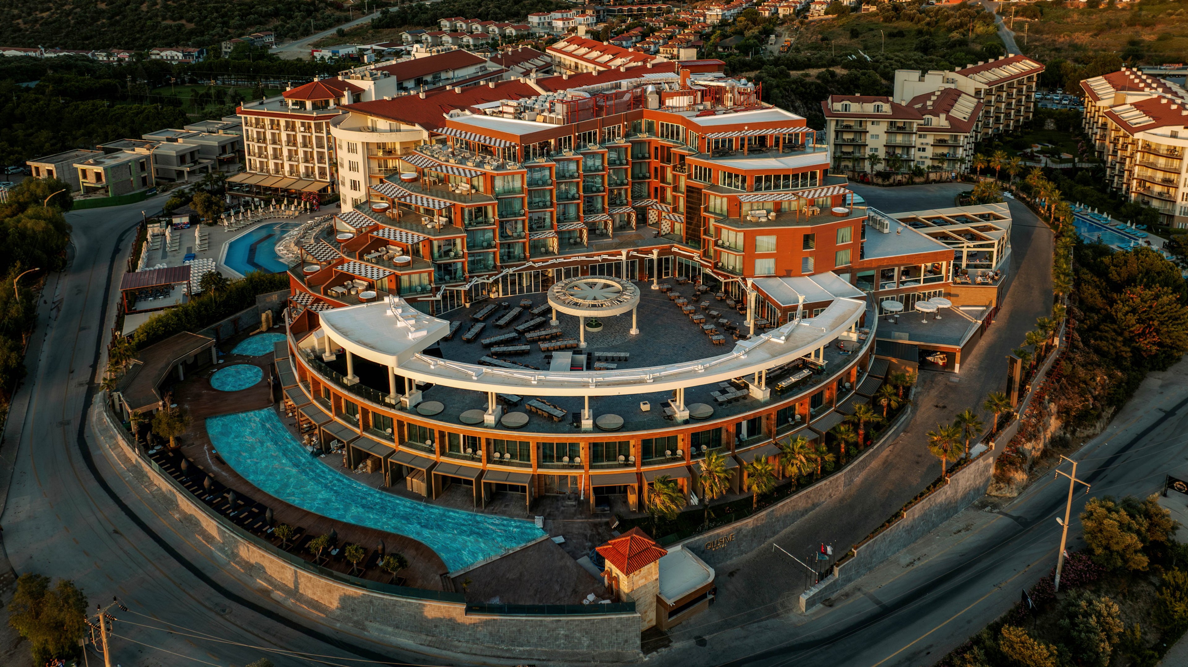 An expansive luxury resort complex is captured from an aerial perspective at golden hour. The multi-story buildings feature red roofs, glass balconies, and a large circular central terrace. Curved swimming pools and landscaped palm-lined walkways surround the structure, emphasizing its resort atmosphere. The property is bordered by winding roads and set against a hilly coastal landscape, suggesting a scenic vacation destination.