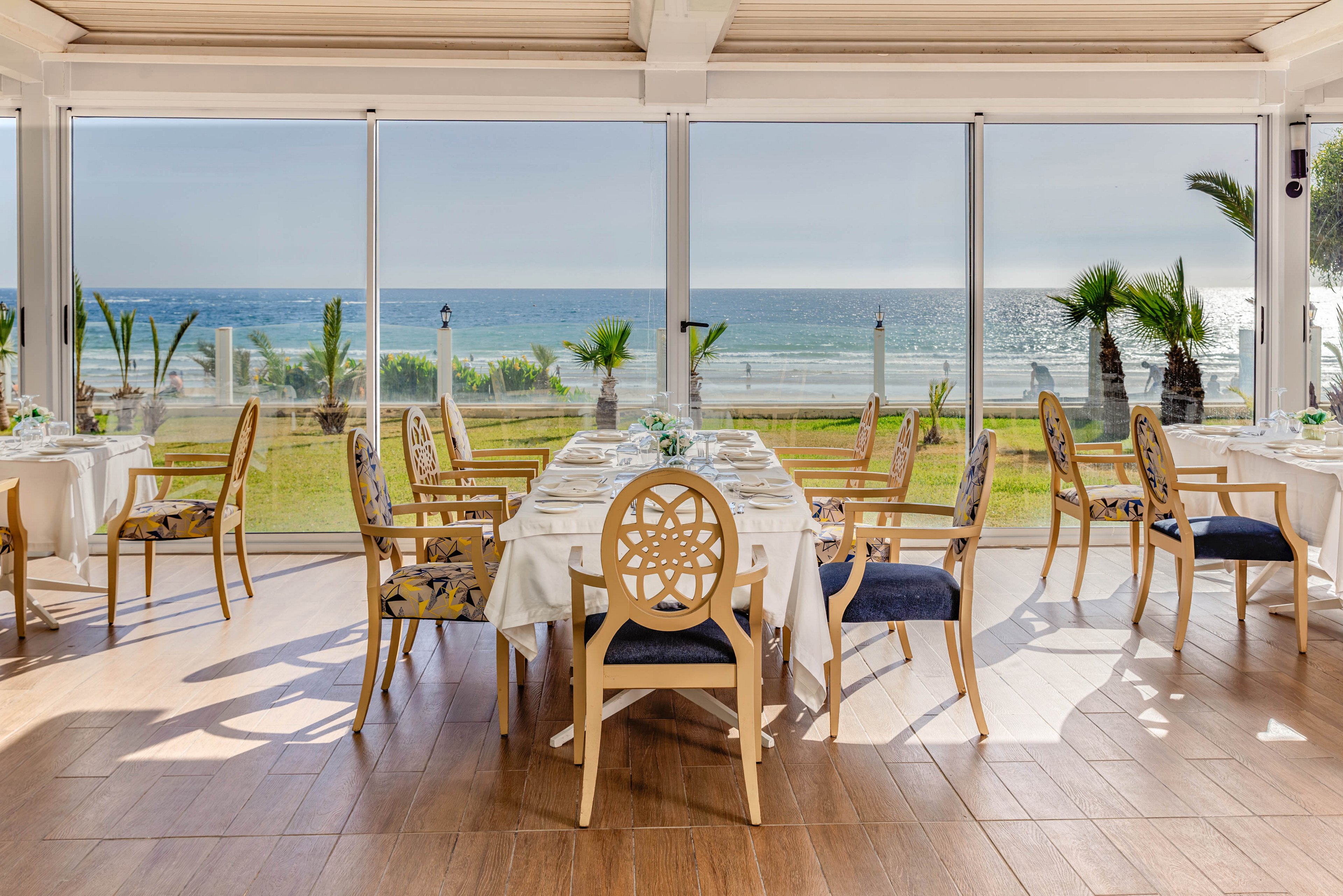 Bright, upscale restaurant dining room overlooks a sunny oceanfront through large floor-to-ceiling windows. Wooden chairs with decorative backs surround neatly arranged tables covered in white tablecloths. The polished wooden floor and neutral interior tones contrast with the green lawn, palm trees, and sparkling blue sea outside. The scene suggests a tranquil, refined coastal dining experience in daylight.