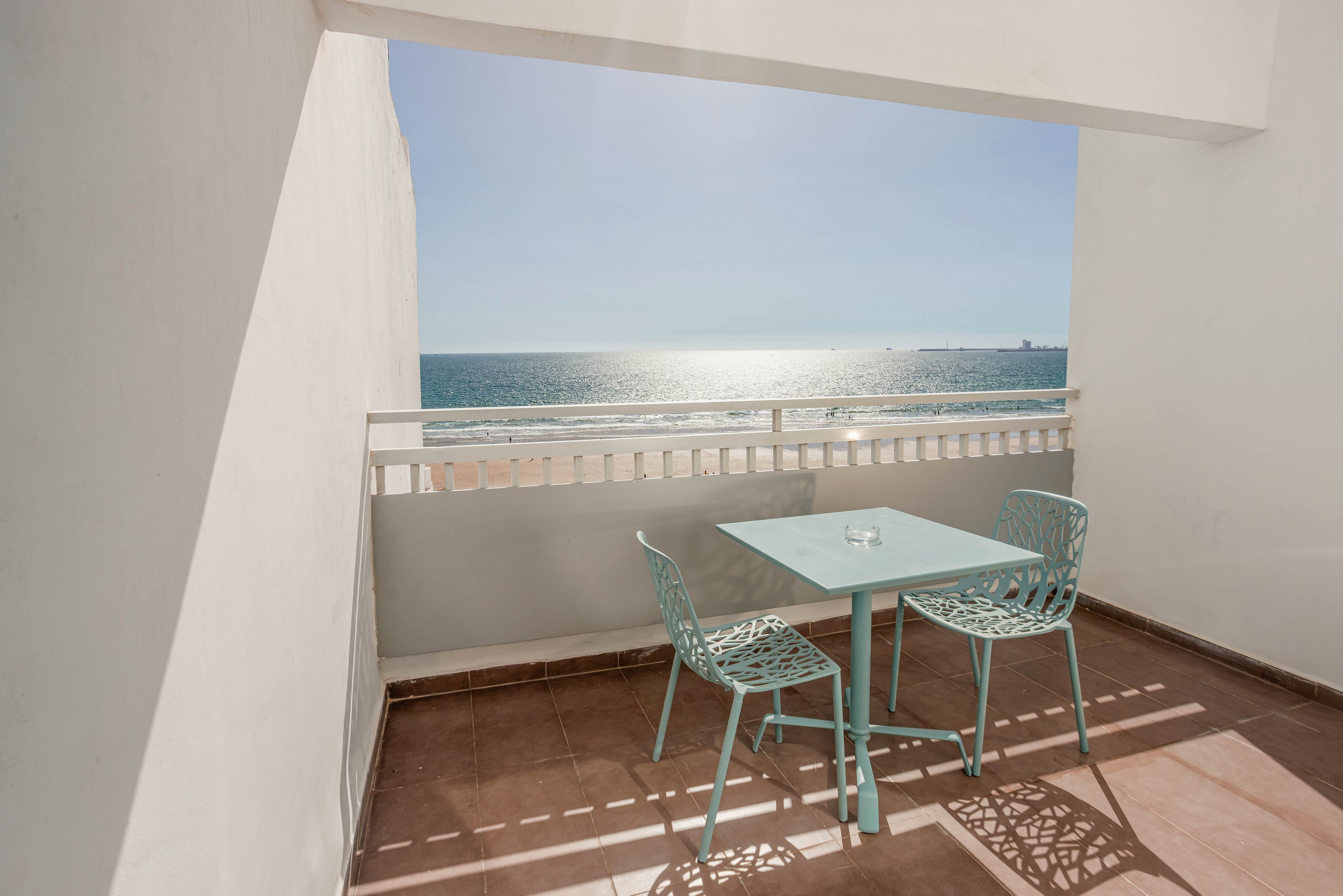 A small balcony terrace features a light blue metal table and two matching chairs. The space is framed by white walls and a railing, opening to a clear view of the shimmering ocean. Bright sunlight casts geometric shadows on the tiled floor, creating a clean and airy atmosphere. The scene evokes a peaceful seaside retreat under a clear blue sky.