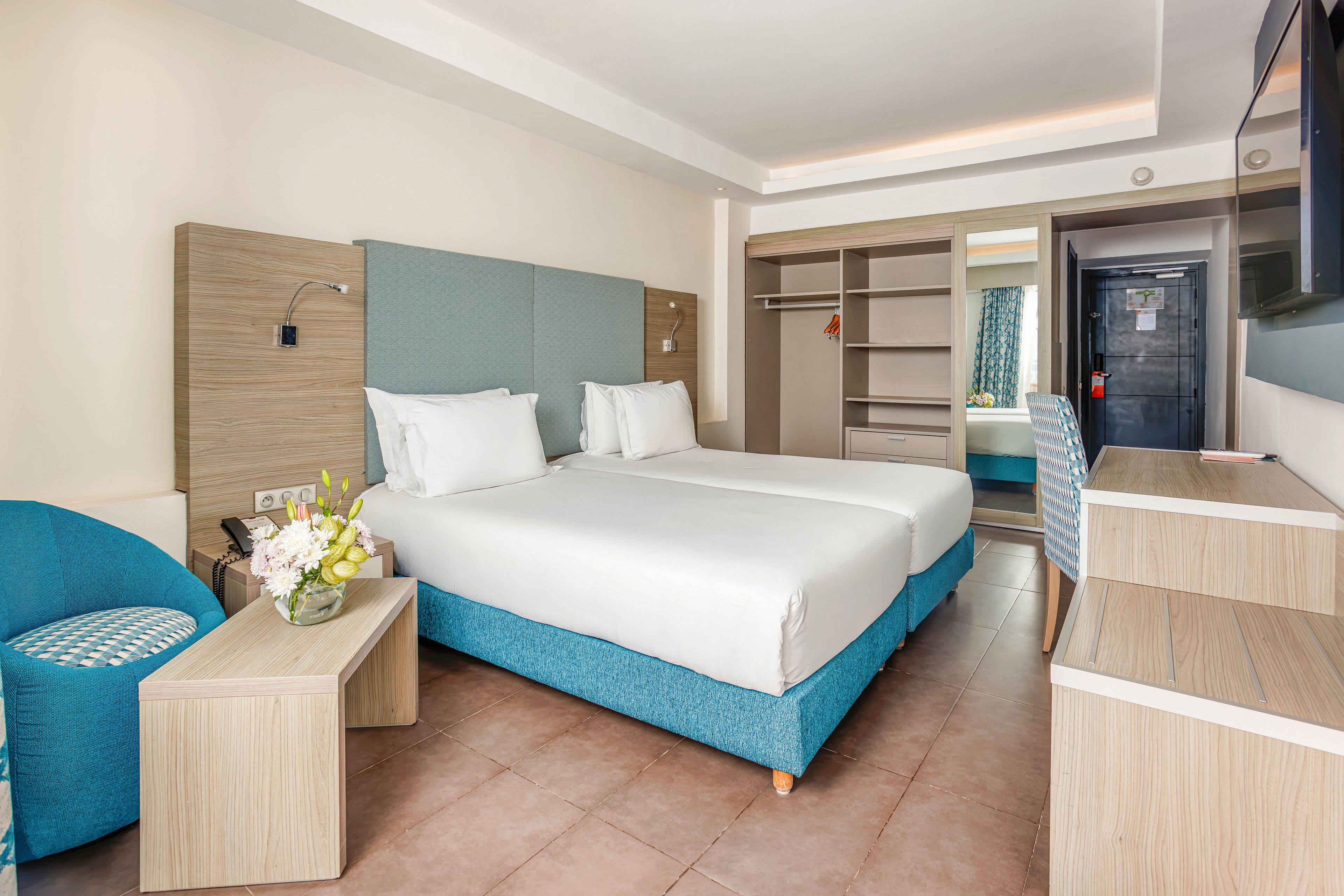 Contemporary hotel room featuring two neatly made twin beds with white linens and teal upholstered bases. The space includes light wood furniture, such as bedside tables, a desk, and open wardrobe shelving. A teal armchair with a patterned cushion and a vase of flowers add color and comfort. Soft lighting and neutral walls create a clean, inviting atmosphere suitable for travelers.