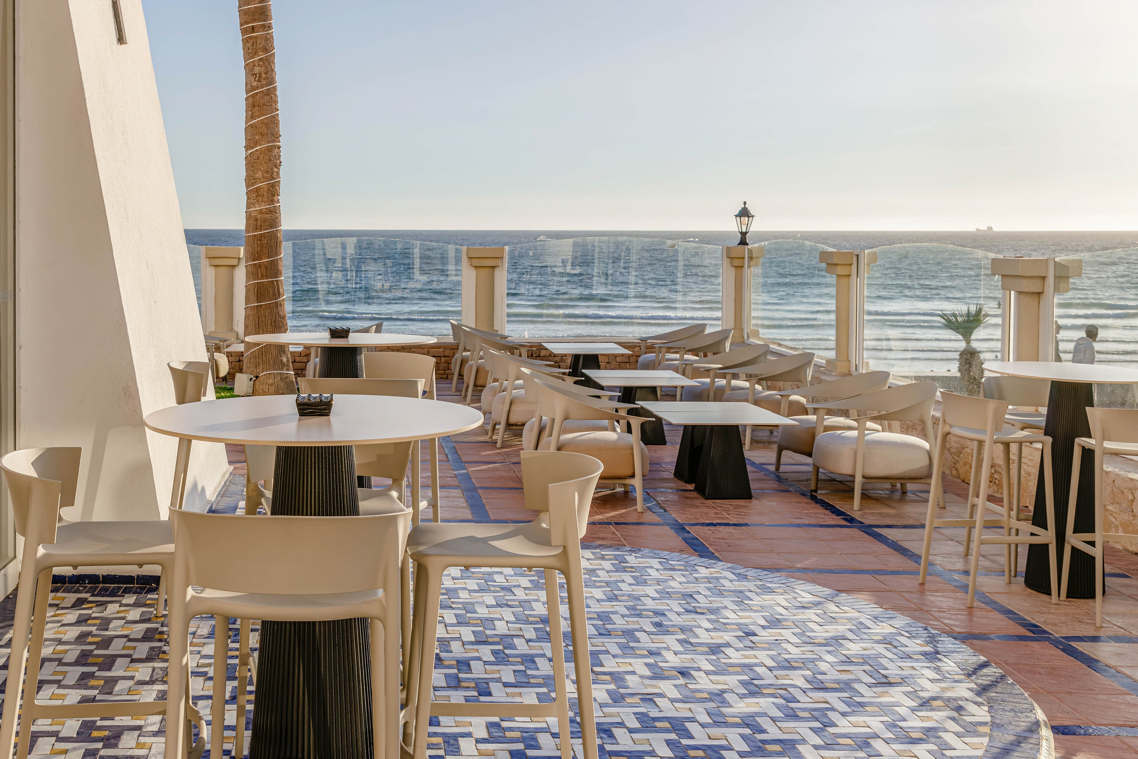 An open-air terrace bar overlooks a calm ocean under clear skies. Light-colored tables and chairs are arranged on a patterned tile floor, facing the water. Tall stools and lounge seating create a relaxed, resort-style atmosphere. Warm sunlight enhances the coastal setting and highlights the tranquil seaside mood.