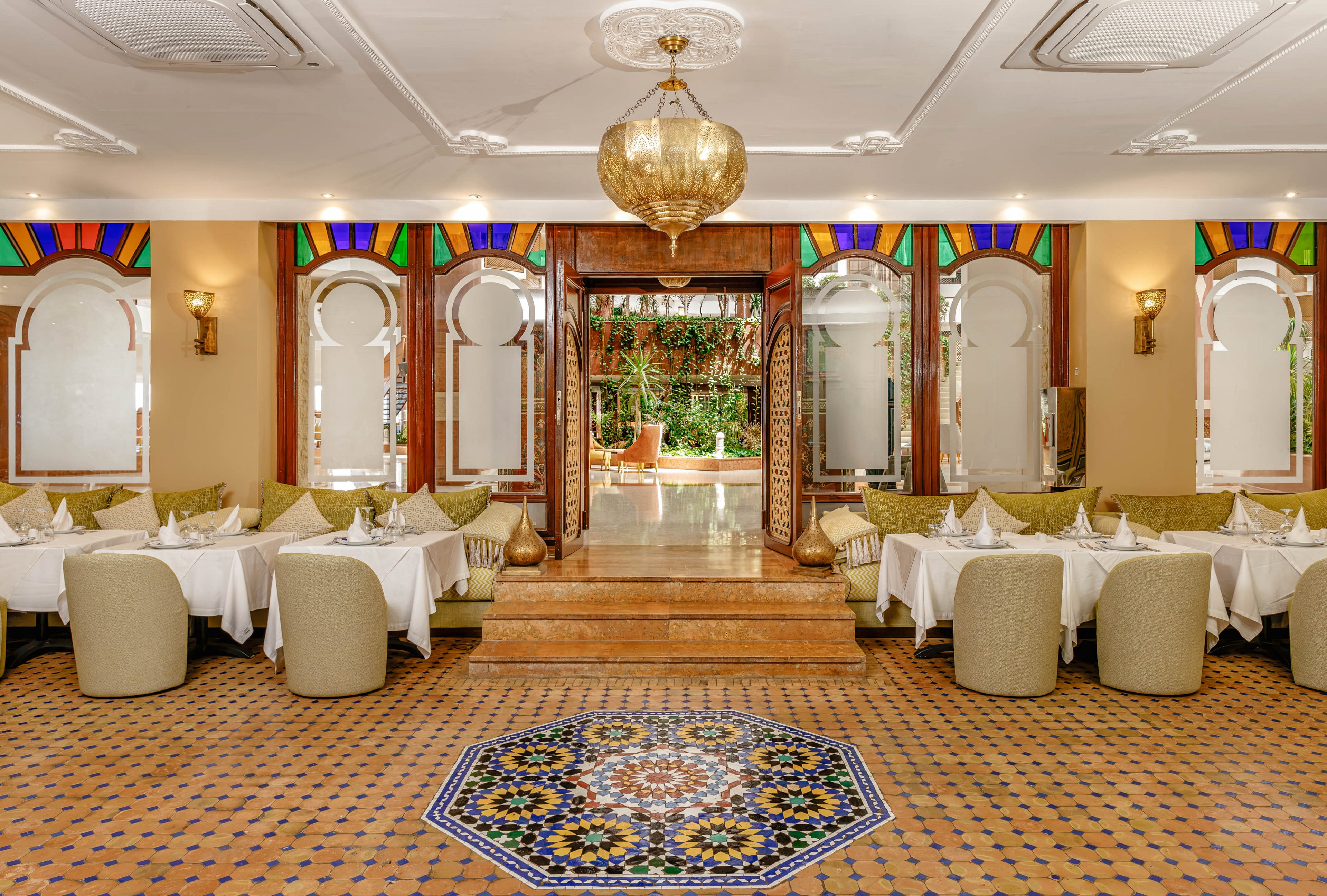 Upscale restaurant dining room featuring white tablecloths and neatly arranged place settings. The space is decorated with colorful stained glass accents above arched mirrors and windows, and a large ornate chandelier hangs from the ceiling. A vibrant geometric mosaic tile pattern anchors the center of the floor, leading toward a raised doorway that opens to a lush garden terrace. The overall ambiance is warm, inviting, and sophisticated, with beige tones complemented by rich jewel colors.