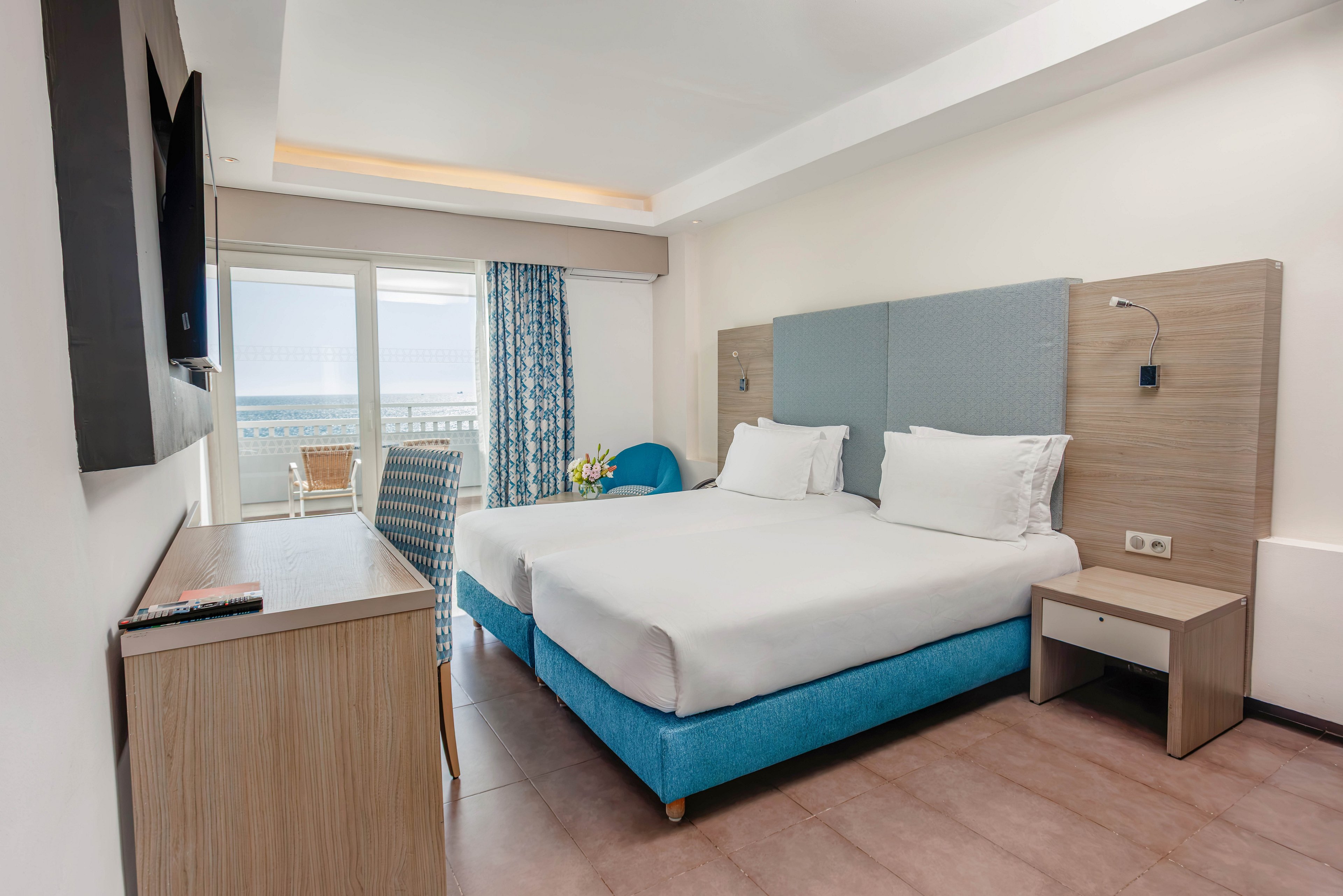Bright contemporary hotel room featuring two neatly made twin beds with blue upholstered bases and light wood headboards. A wall-mounted television faces the beds above a simple wooden desk and chair. Large glass doors open to a balcony with outdoor seating and a clear view of the ocean. Soft neutral tones are accented by blue fabrics and patterned curtains, creating a clean and relaxing atmosphere.
