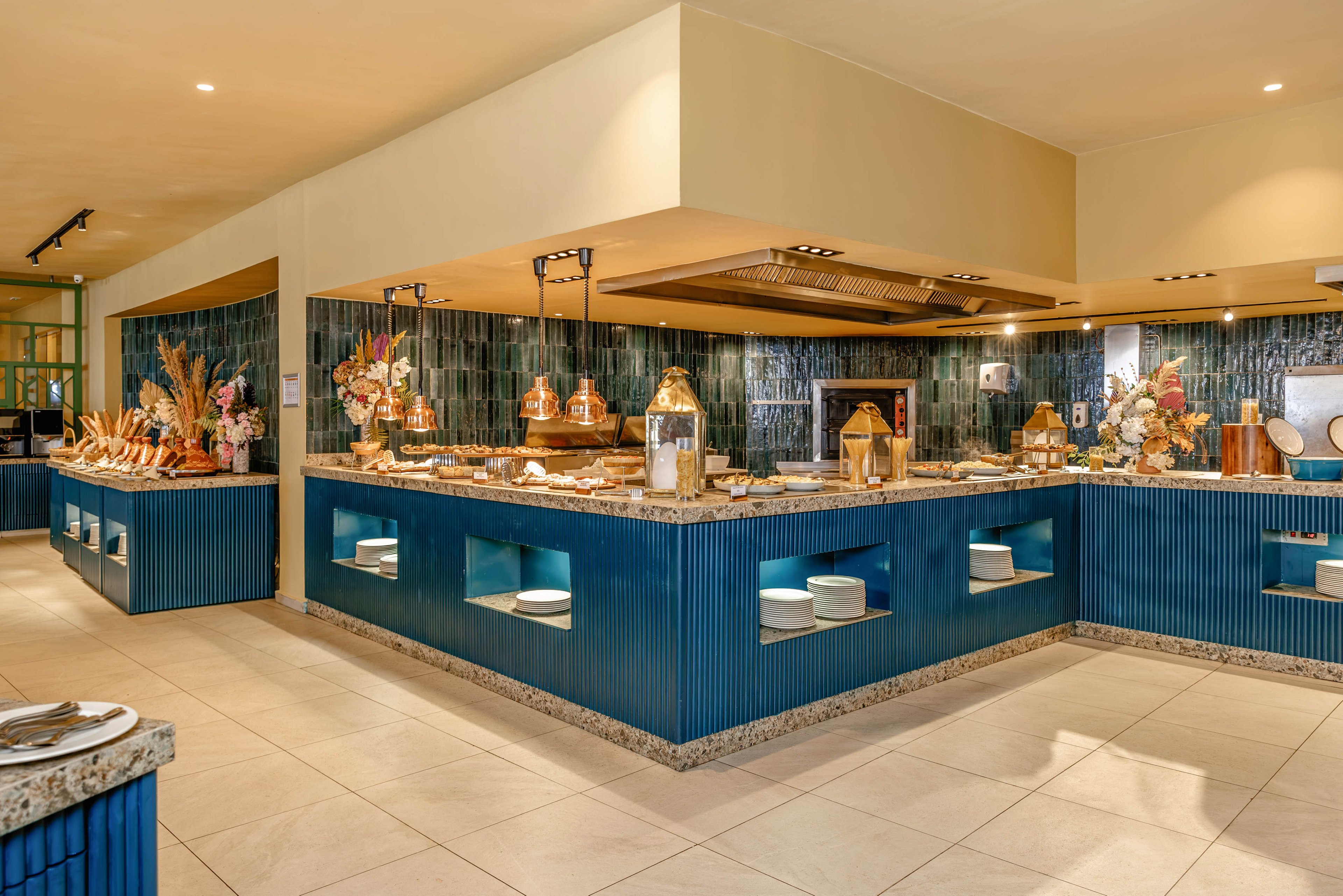 A spacious buffet area features blue ribbed counters with stone countertops arranged in a U-shaped layout. Various serving dishes, decorative vases, and dried floral arrangements are displayed under warm overhead lighting. The background includes tiled walls, open shelving with plates, and neatly organized food presentation areas. The overall setting feels modern, clean, and inviting, suitable for a hotel or resort restaurant.