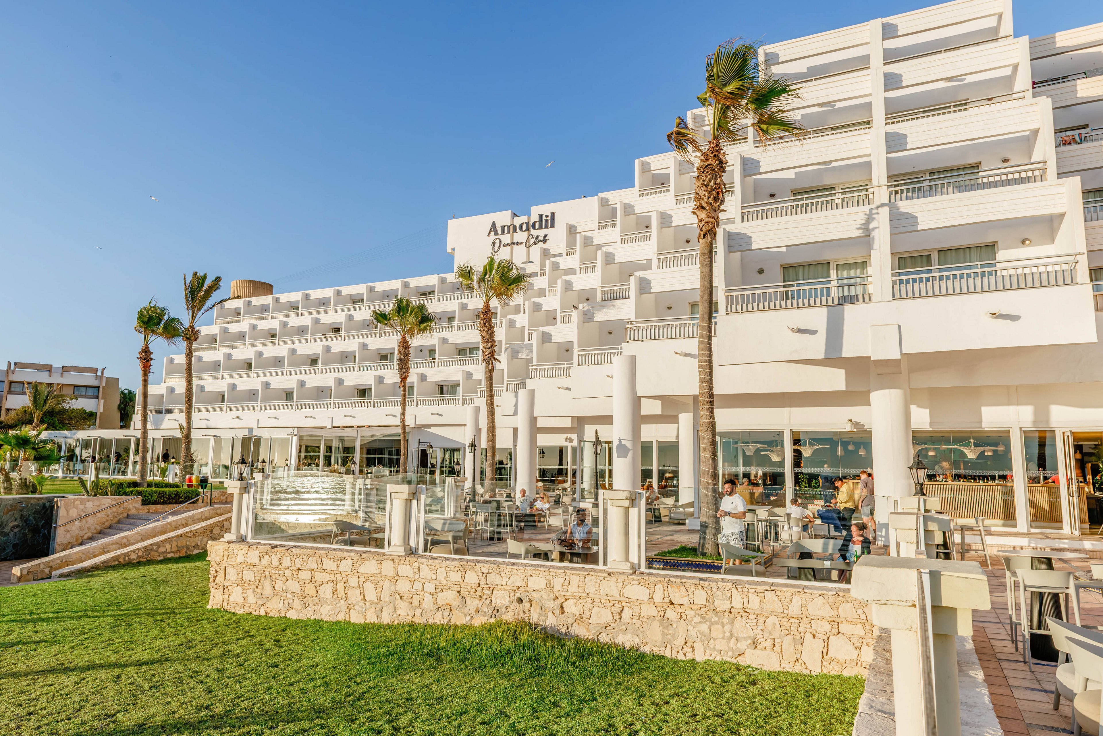 A large white multi-story hotel building stands beside a green lawn and outdoor terrace area. Guests sit at tables under clear skies, enjoying the open-air restaurant and bar. Tall palm trees line the front of the hotel, adding a tropical feel to the coastal setting. Glass railings, stone walls, and bright sunlight create a clean, contemporary atmosphere.
