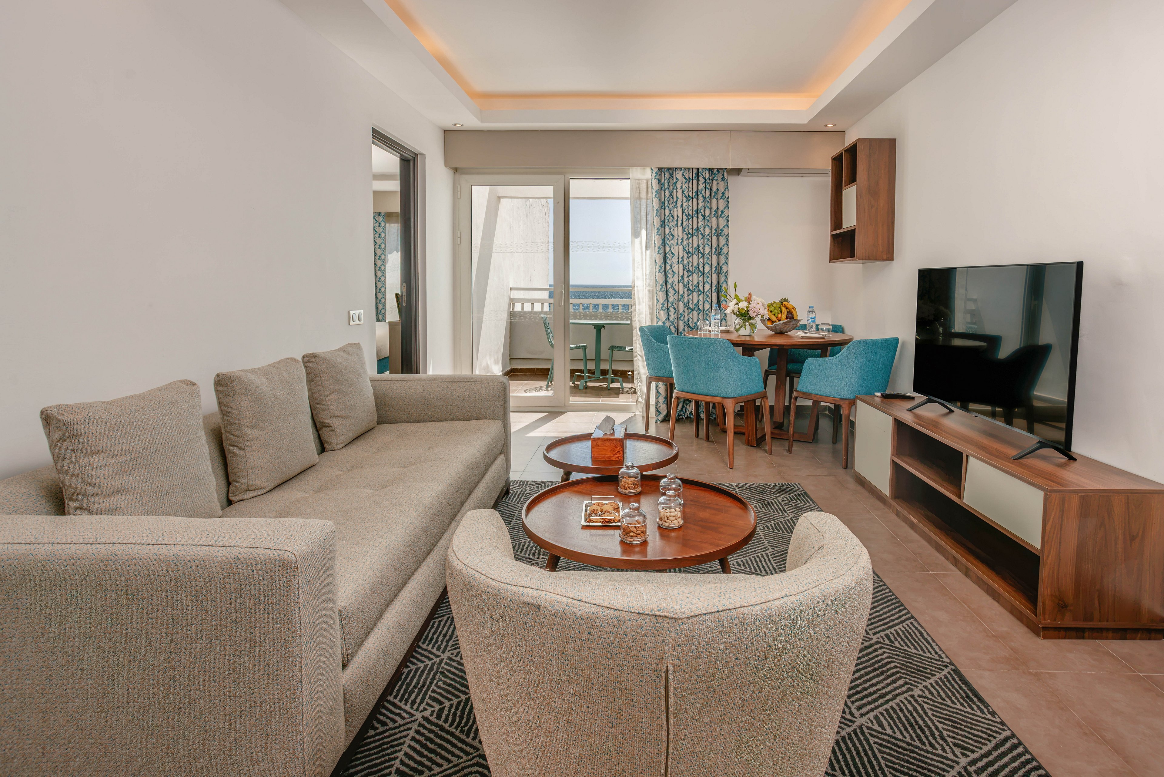 Contemporary hotel suite living area with a beige sofa and armchairs faces a flat-screen television on a wooden media unit. Two round coffee tables hold glasses and decorative items, while a patterned rug defines the seating space. In the background, a dining table with blue chairs is set with flowers and tableware near large glass doors. The doors open to a balcony with a view of the sea, bringing natural light into the bright, neutral-toned room.