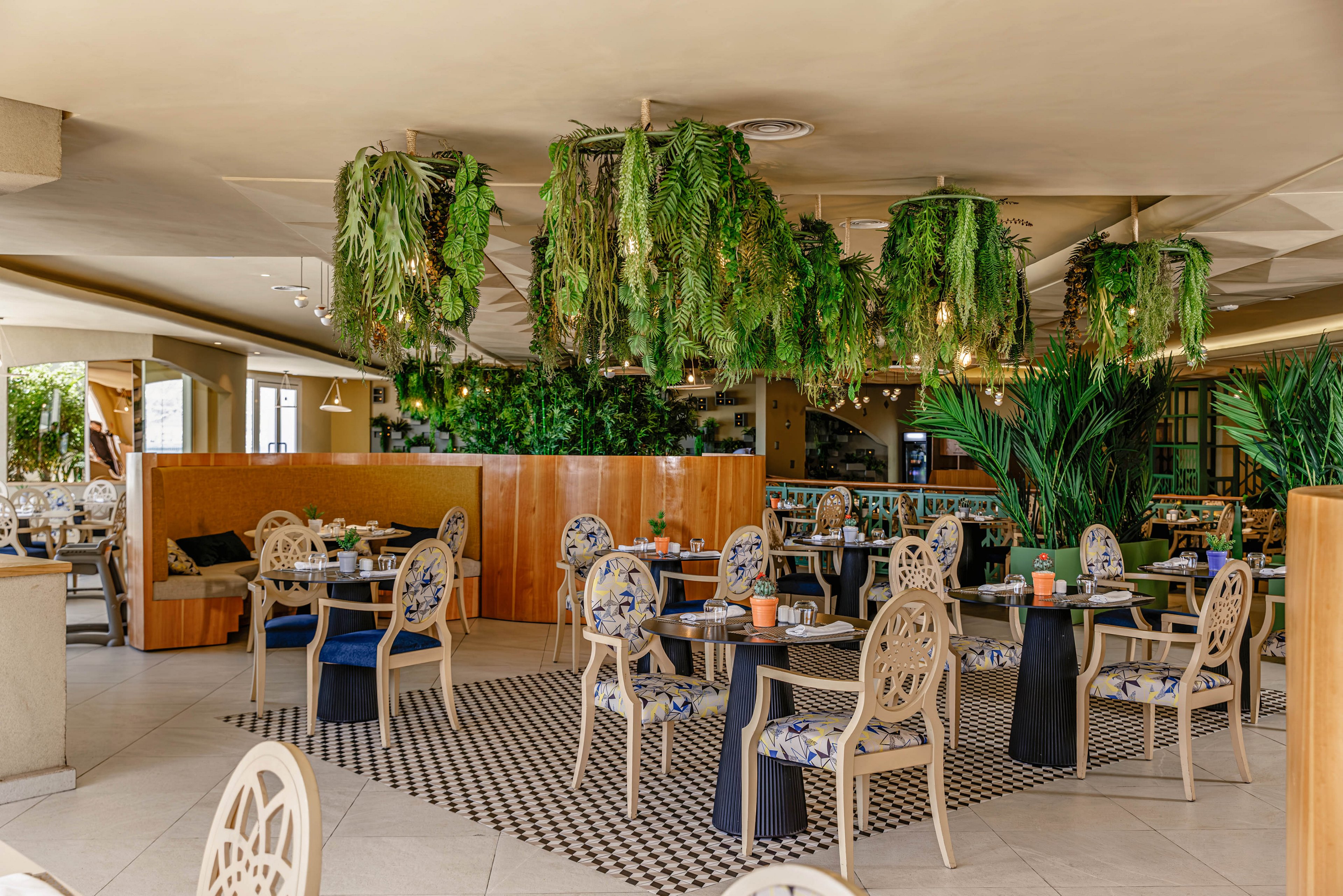 Contemporary restaurant dining area featuring elegant chairs and neatly set tables. The space is decorated with abundant hanging plants and large potted greenery, creating a fresh, natural atmosphere. Warm wood paneling and patterned floor tiles add visual interest and contrast with the neutral walls. Daylight streams in from large openings, highlighting the relaxed yet upscale setting.