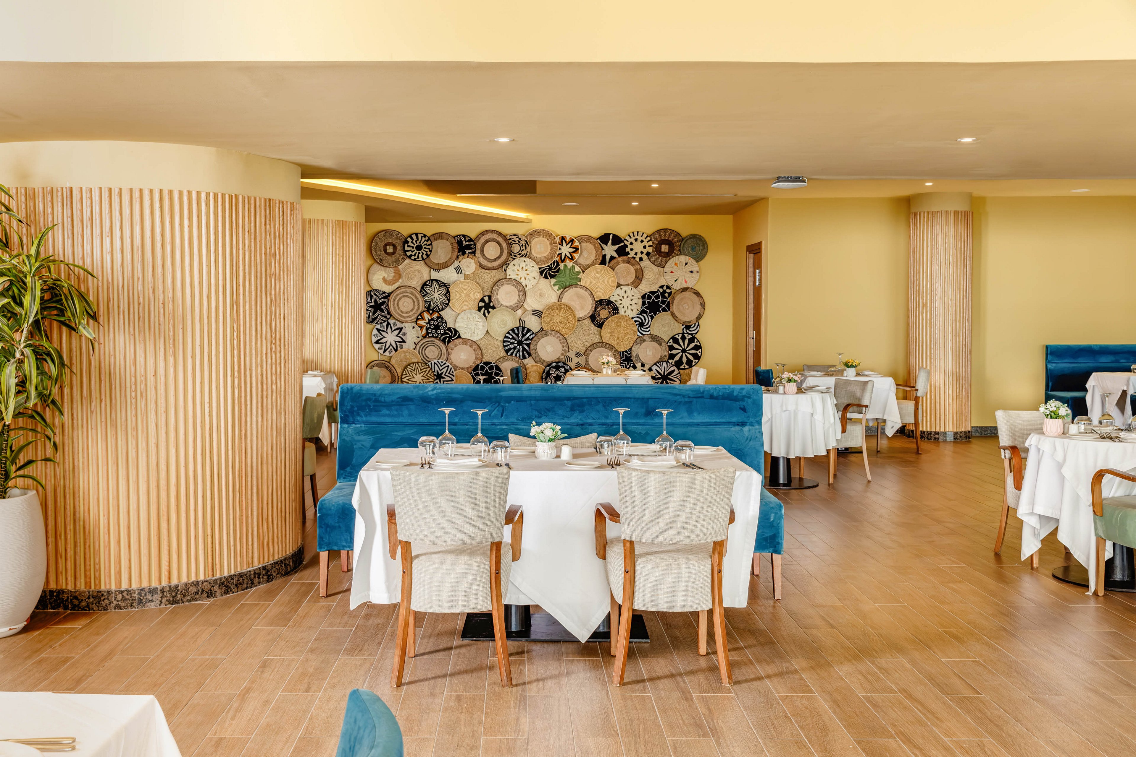 Bright contemporary restaurant interior featuring neatly set tables with white tablecloths and upholstered chairs. A long blue banquette runs along the wall, facing round tables arranged for guests. The back wall is decorated with a large artistic display of circular woven patterns in neutral tones. Warm wood flooring and columns, soft yellow walls, and potted plants create a welcoming atmosphere. Glassware and small floral centerpieces add an elegant finishing touch to the space.
