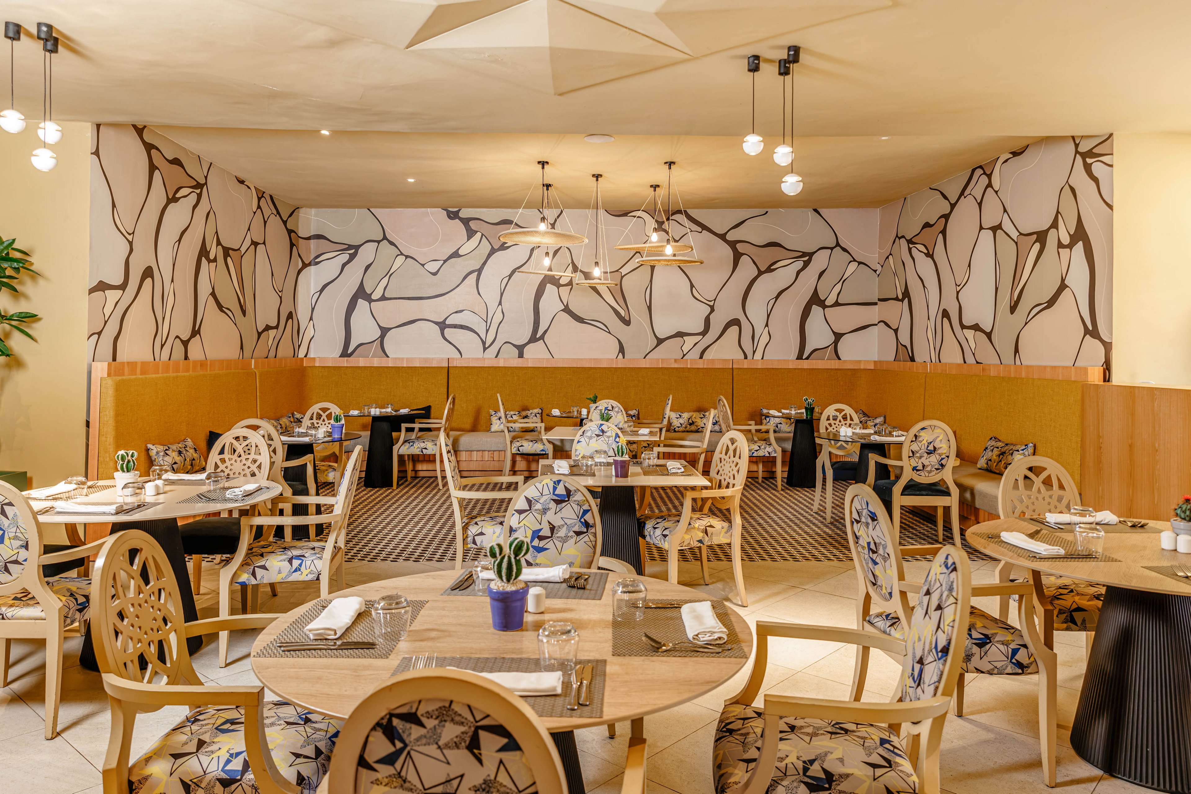 A contemporary restaurant dining room features round wooden tables and decorative chairs arranged neatly across the space. The setting is bright and inviting, with patterned upholstery and a bold abstract mural covering the back wall. Warm pendant lighting hangs from the ceiling, enhancing the cozy atmosphere and highlighting the artistic design elements. Table settings with napkins, plates, and small plants suggest the room is prepared for guests.