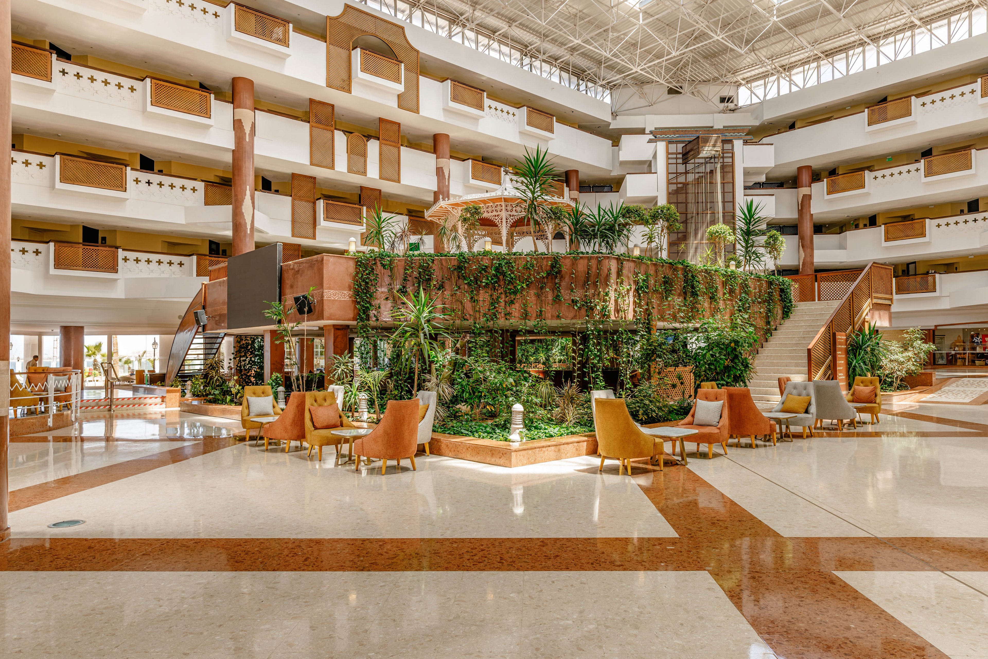 Large open hotel atrium features a central indoor garden surrounded by seating areas. Multiple floors of balconies overlook the bright lobby, illuminated by a high glass ceiling. Green plants cascade over the raised garden structure, contrasting with warm beige and brown architectural details. Comfortable armchairs and tables create a relaxed social space in the middle of the polished floor.