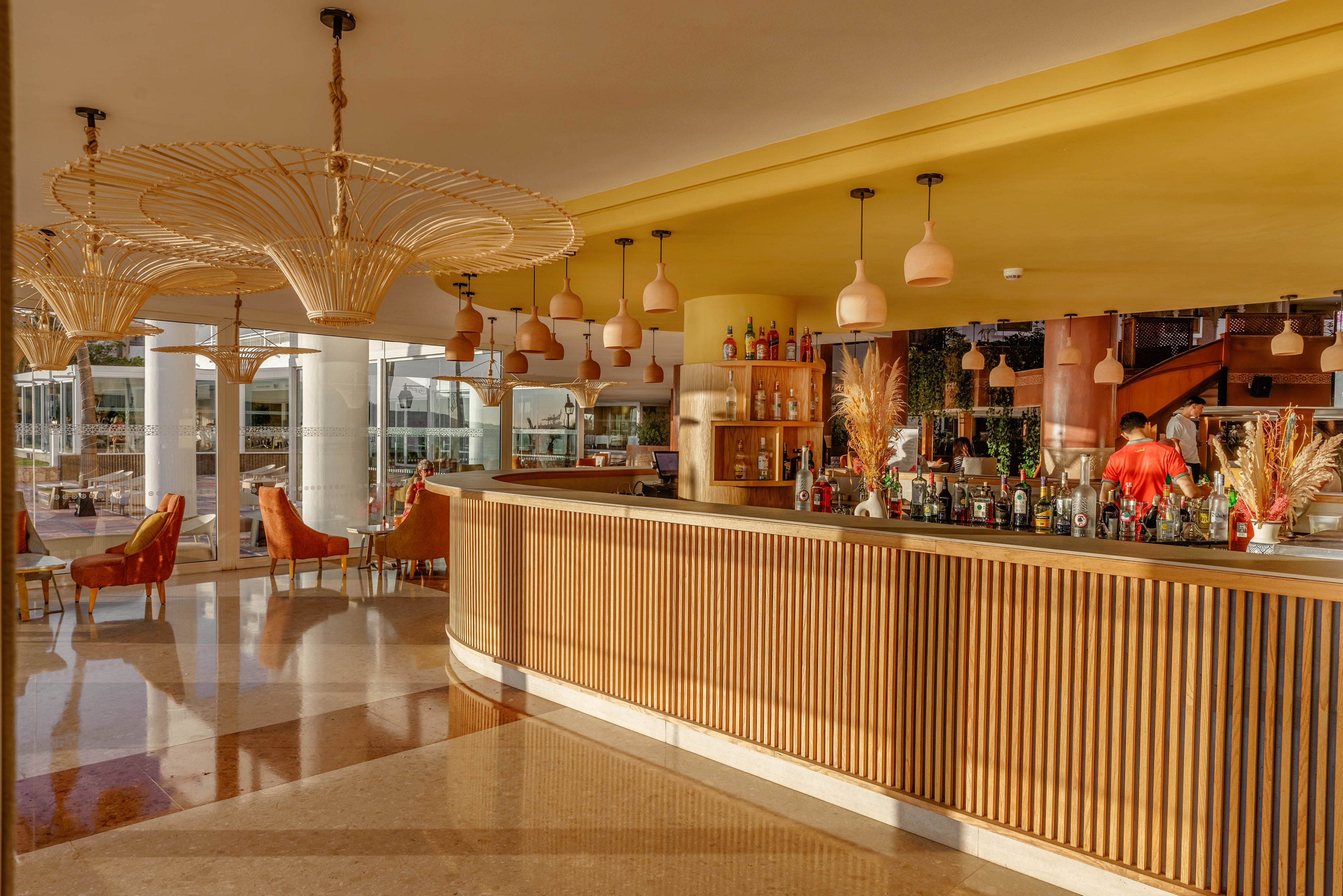 A stylish hotel bar interior features a long wooden counter lined with various bottles and glassware. Warm sunlight streams through large windows, illuminating contemporary pendant lights and wicker-style chairs. A bartender in a red shirt works behind the bar, while seating areas with cushioned armchairs create a relaxed atmosphere. The overall scene combines modern design with a cozy, golden-toned ambiance.