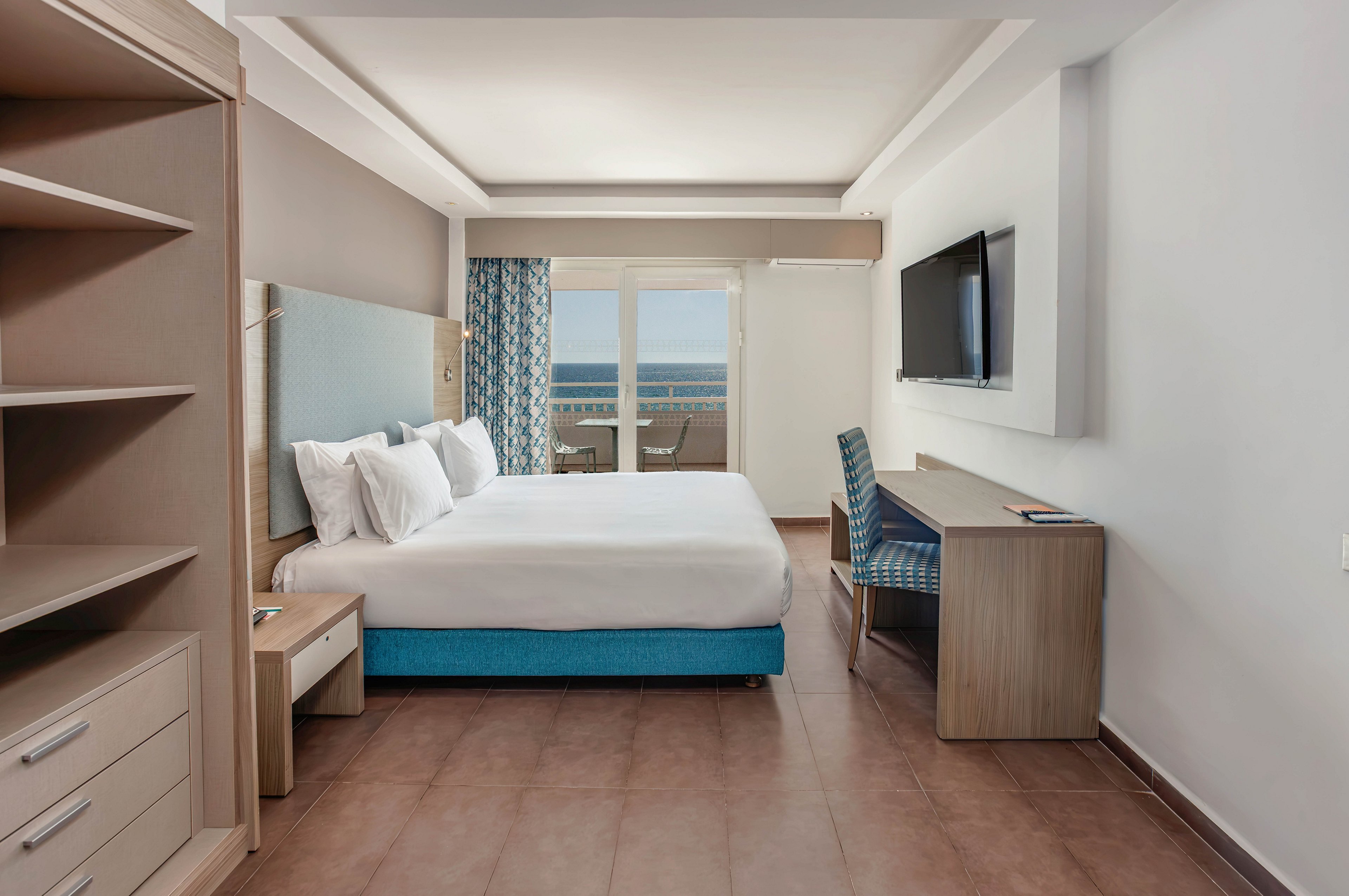 Contemporary hotel room featuring a large double bed with white linens and turquoise base. A wall-mounted flat-screen TV faces the bed above a wooden desk and blue patterned chair. Built-in shelving and drawers line one side of the room, while floor-to-ceiling curtains frame glass doors leading to a balcony. Through the doors, a clear view of the sea and horizon enhances the bright, minimalist interior.