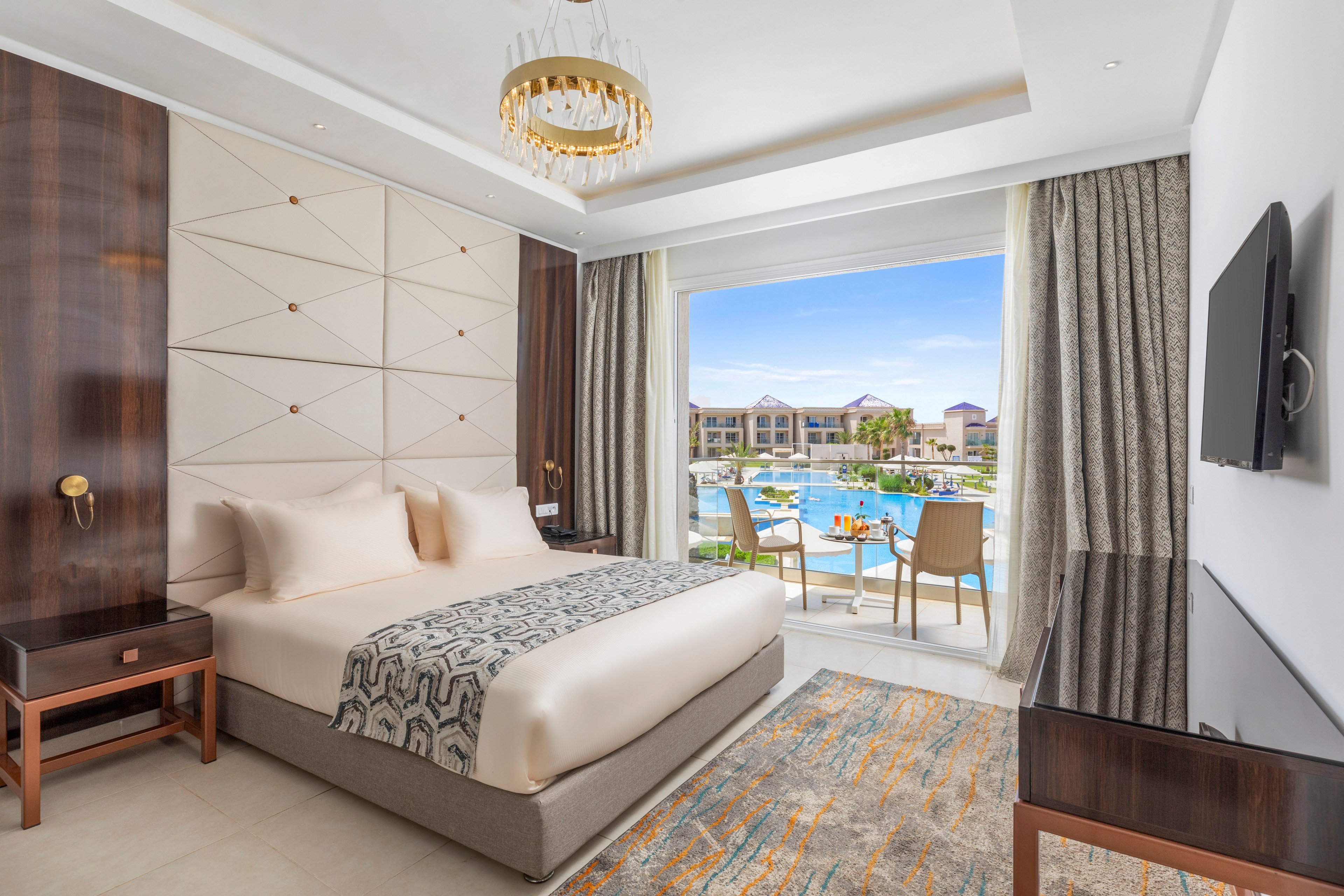 A contemporary hotel room features a neatly made double bed with neutral bedding and a patterned throw. The interior includes sleek wooden furniture, a wall-mounted television, and a decorative chandelier. Large curtains frame sliding glass doors that open to a balcony overlooking a resort pool and surrounding buildings. Bright daylight and soft tones create a clean, inviting atmosphere ideal for vacation or business travel.