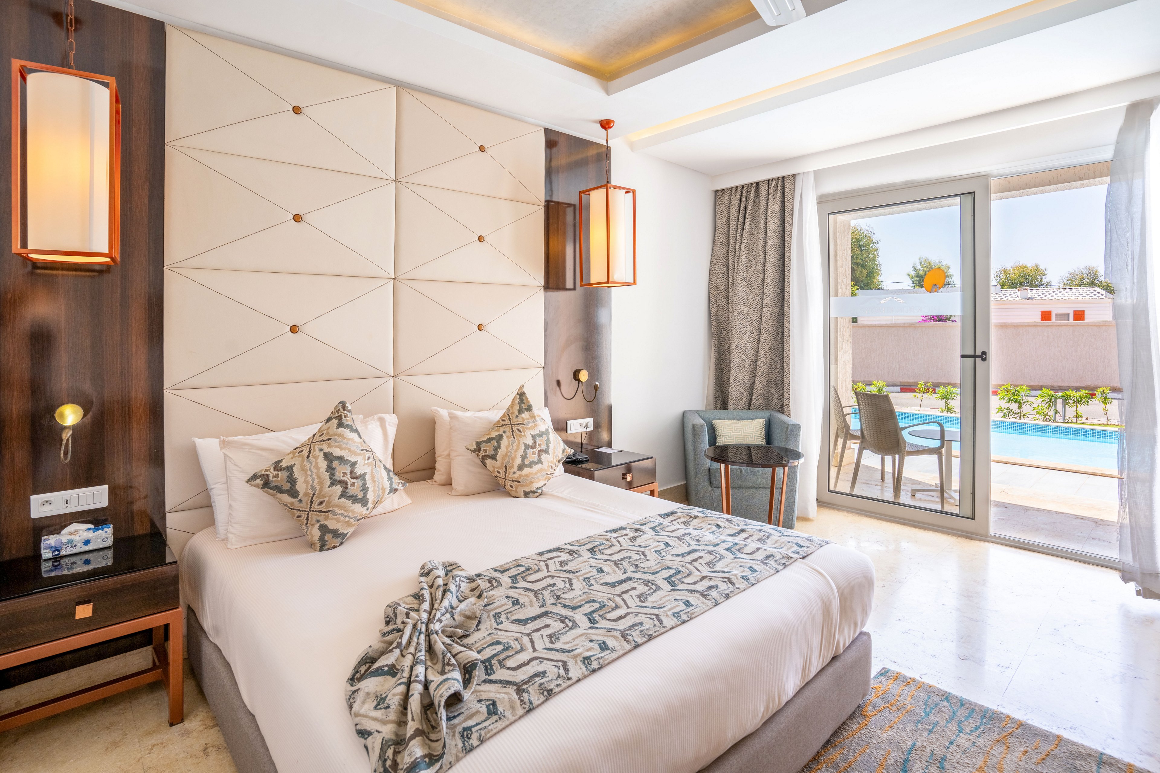 Contemporary hotel room featuring a neatly made double bed with patterned pillows and throw. The space includes warm wooden accents, geometric wall paneling, and soft ambient lighting. A small table and chairs sit beside a large glass door leading to a balcony overlooking a swimming pool and greenery. Natural daylight fills the room, creating an inviting and relaxing atmosphere.