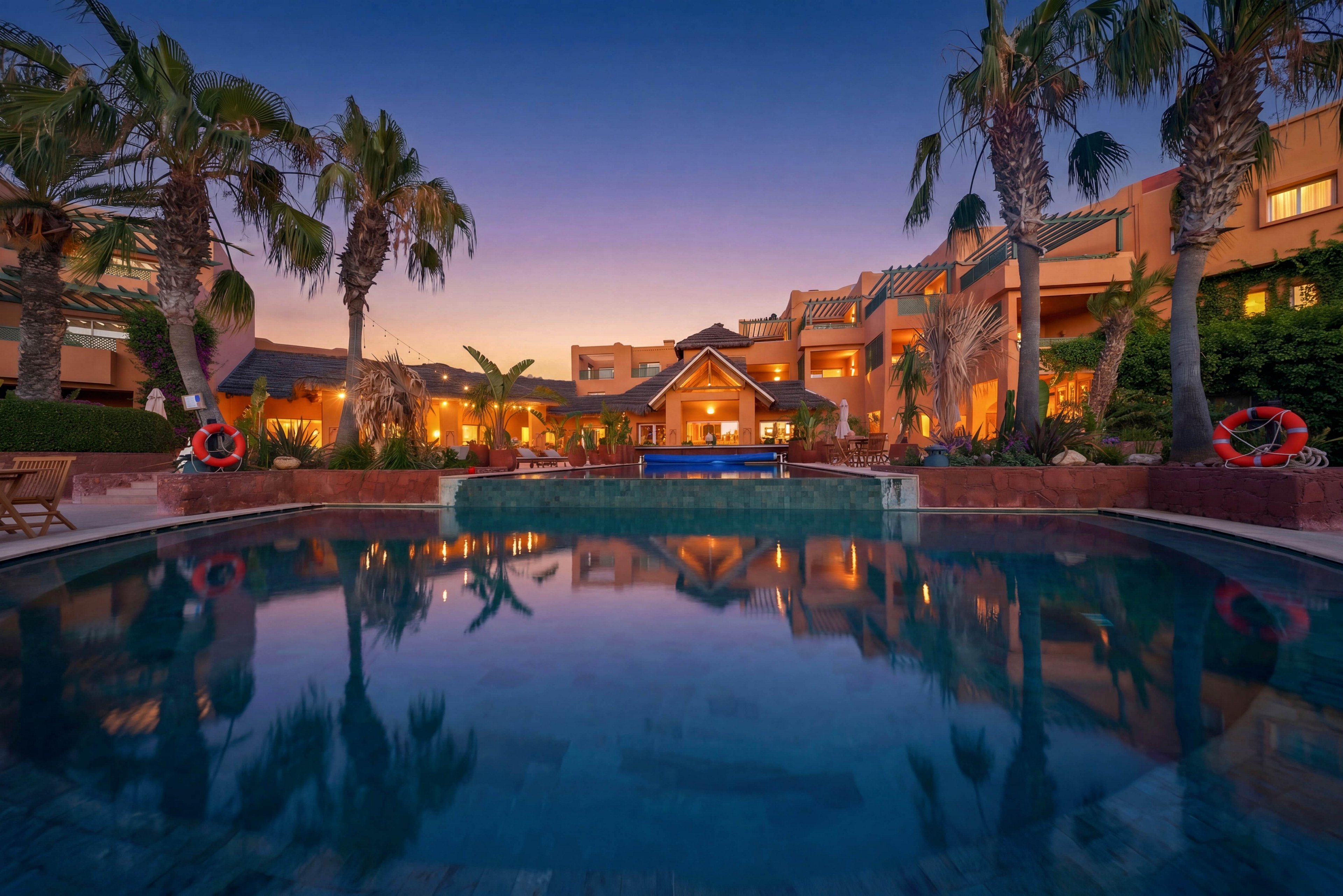 A tranquil resort swimming pool reflects the warm lights of surrounding buildings at dusk. Tall palm trees frame the scene, creating a tropical atmosphere around the water. The sky transitions from deep blue to soft orange, enhancing the serene mood. Poolside safety rings and subtle landscaping details add to the impression of a well-maintained vacation destination.