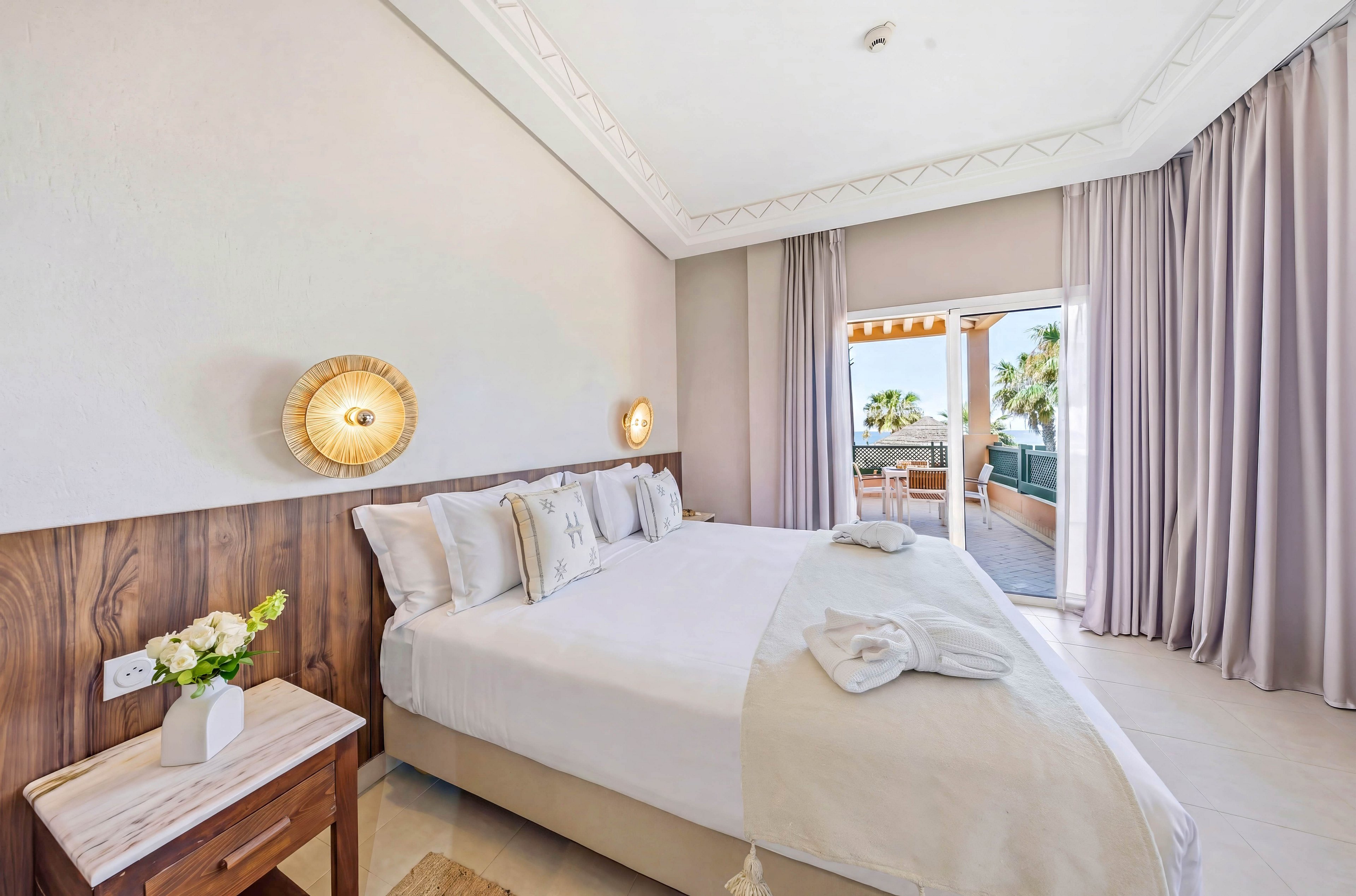 A bright, modern hotel bedroom features a large neatly made bed with white linens and decorative pillows. Warm wooden accents and soft beige tones create a calm, coastal atmosphere. Wall-mounted lights, a bedside table with flowers, and floor-to-ceiling curtains add a touch of elegance. Sliding glass doors open to a furnished balcony with outdoor seating and palm trees visible in the background.