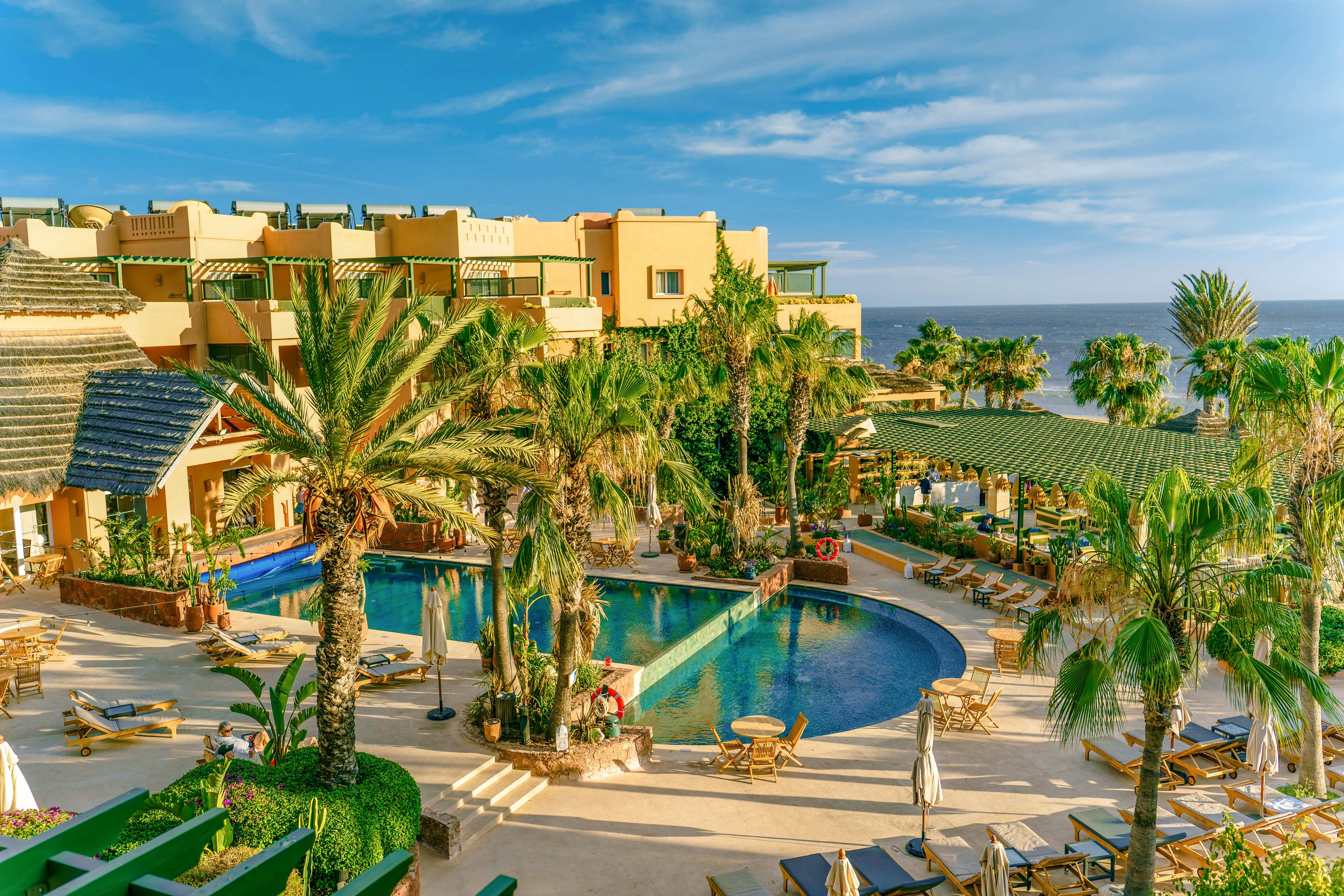 A sunny seaside resort features a large circular swimming pool surrounded by palm trees and sun loungers. Multi-story hotel buildings with balconies overlook the pool area, creating a vibrant vacation atmosphere. Guests relax on deck chairs and walk along the pathways under clear blue skies. The ocean is visible in the background, enhancing the tropical holiday setting.