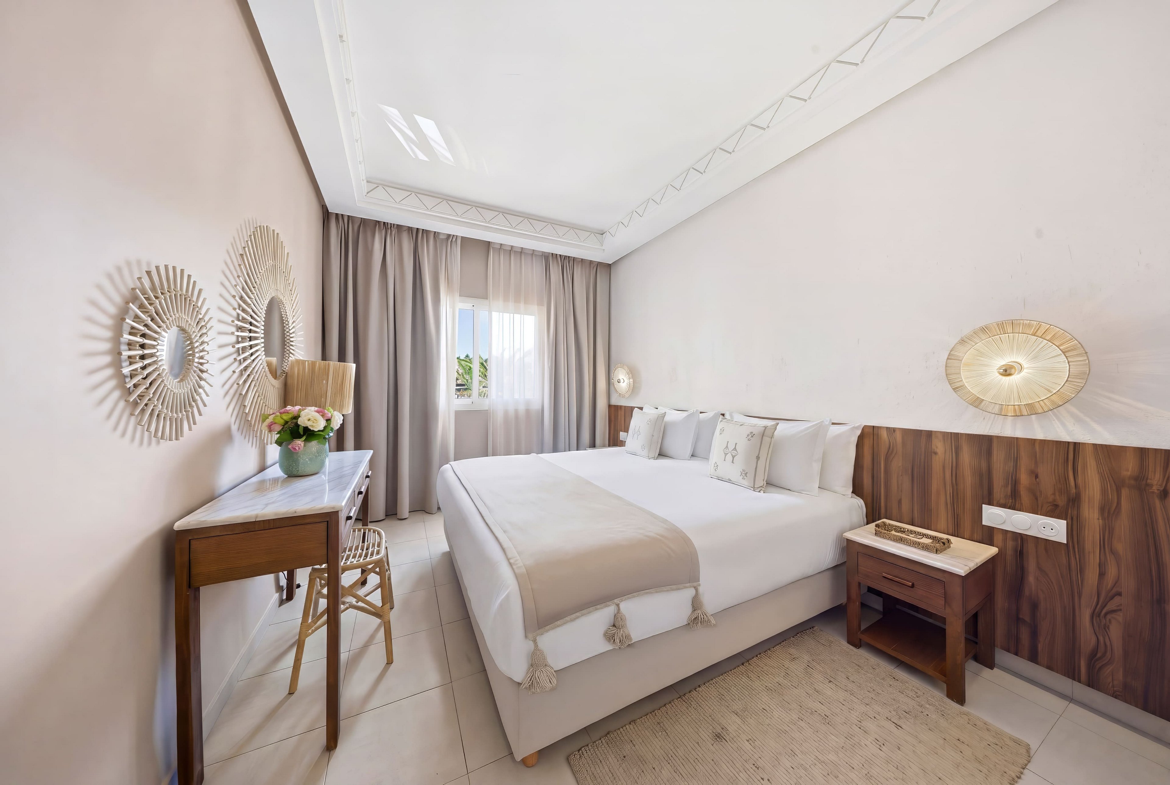 A bright, neatly made double bed dominates a minimalist hotel-style bedroom. The room features soft beige and white tones, with a wooden headboard, bedside table, and a small writing desk with a chair. Decorative round wall mirrors and woven wall lights add texture and interest to the neutral palette. Natural light enters through a curtained window, creating a calm and inviting atmosphere.